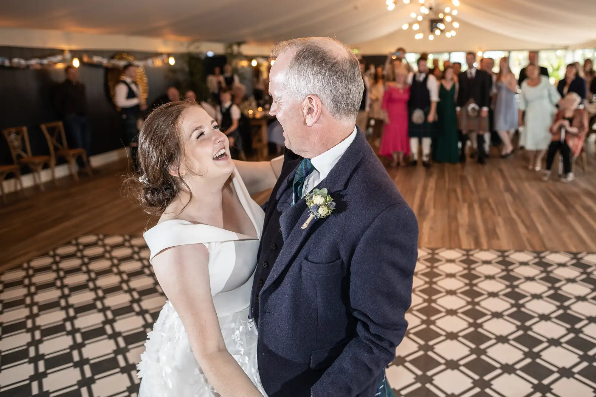 A bride and an older man dance and smile at an indoor wedding reception with guests watching in the background.