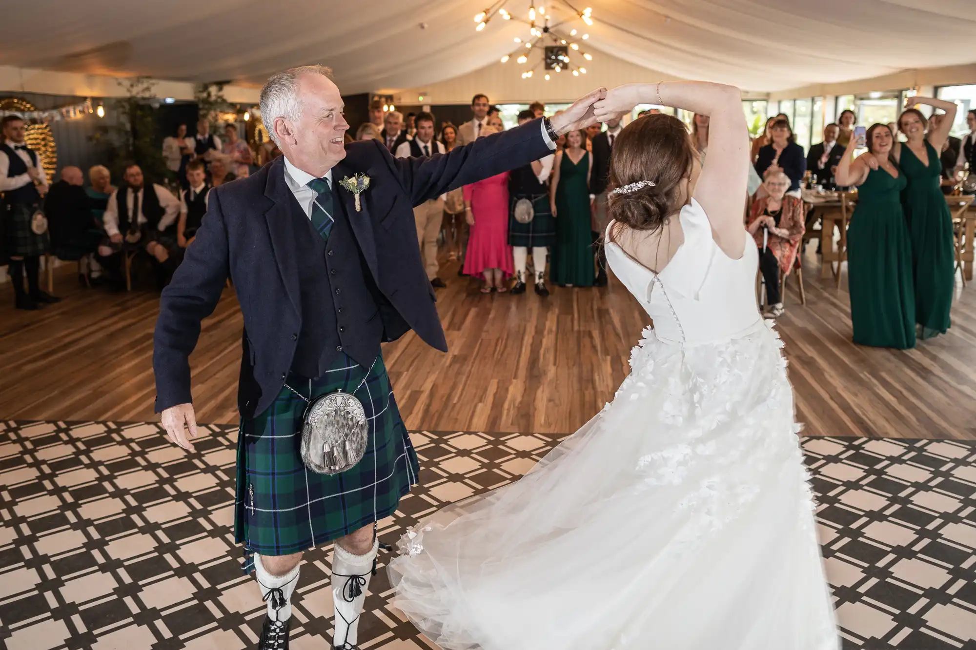 An older man in a kilt dances with a bride in a white dress at a wedding reception. Guests in formal attire watch and cheer in the background.