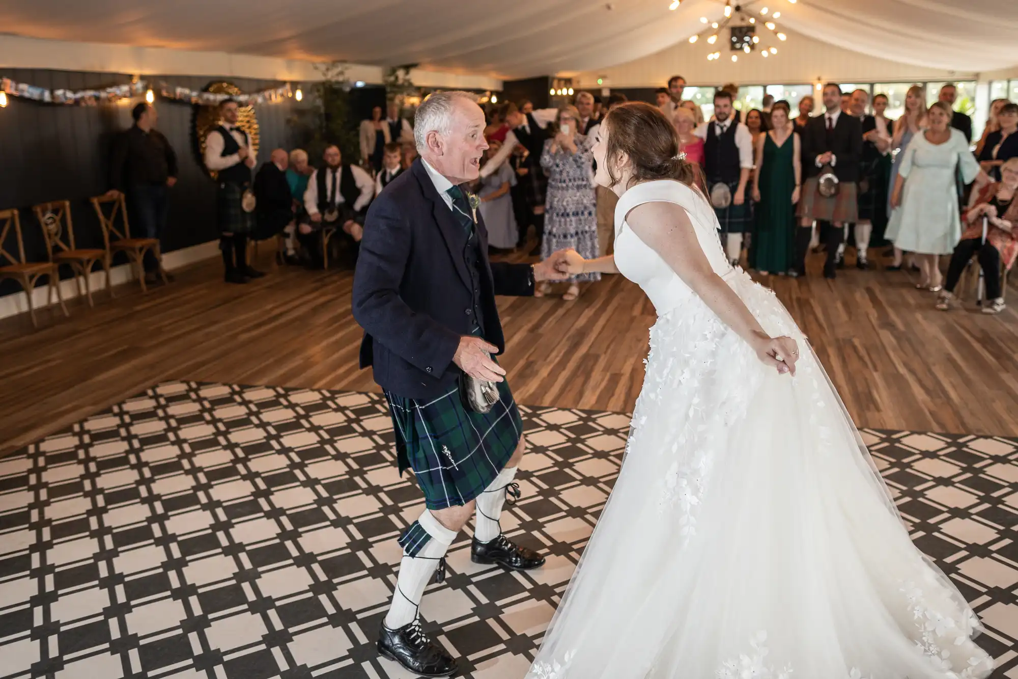 An elderly man in a kilt and a bride in a white gown are dancing joyfully on a checkered floor at a wedding reception, with guests watching in the background.