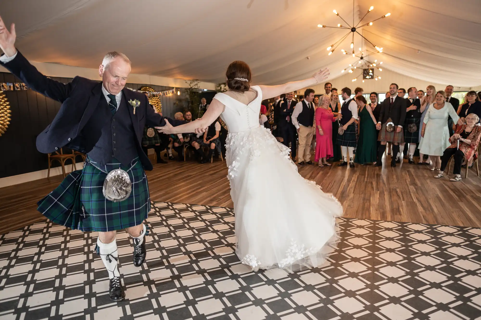 A man in a kilt and a woman in a white dress dance energetically on a checkered floor while a group of people watches under a tent with string lights.