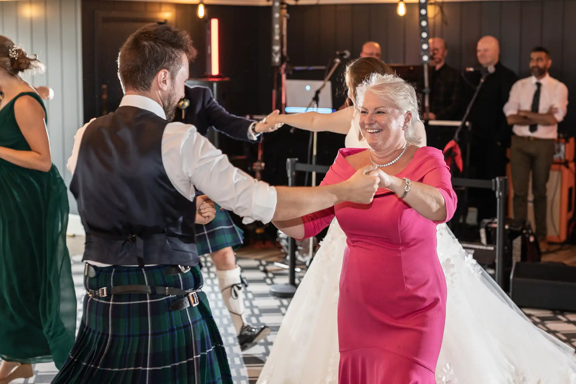 People dancing at an event, with one man in a kilt and a woman in a pink dress in the foreground. A band is playing in the background.