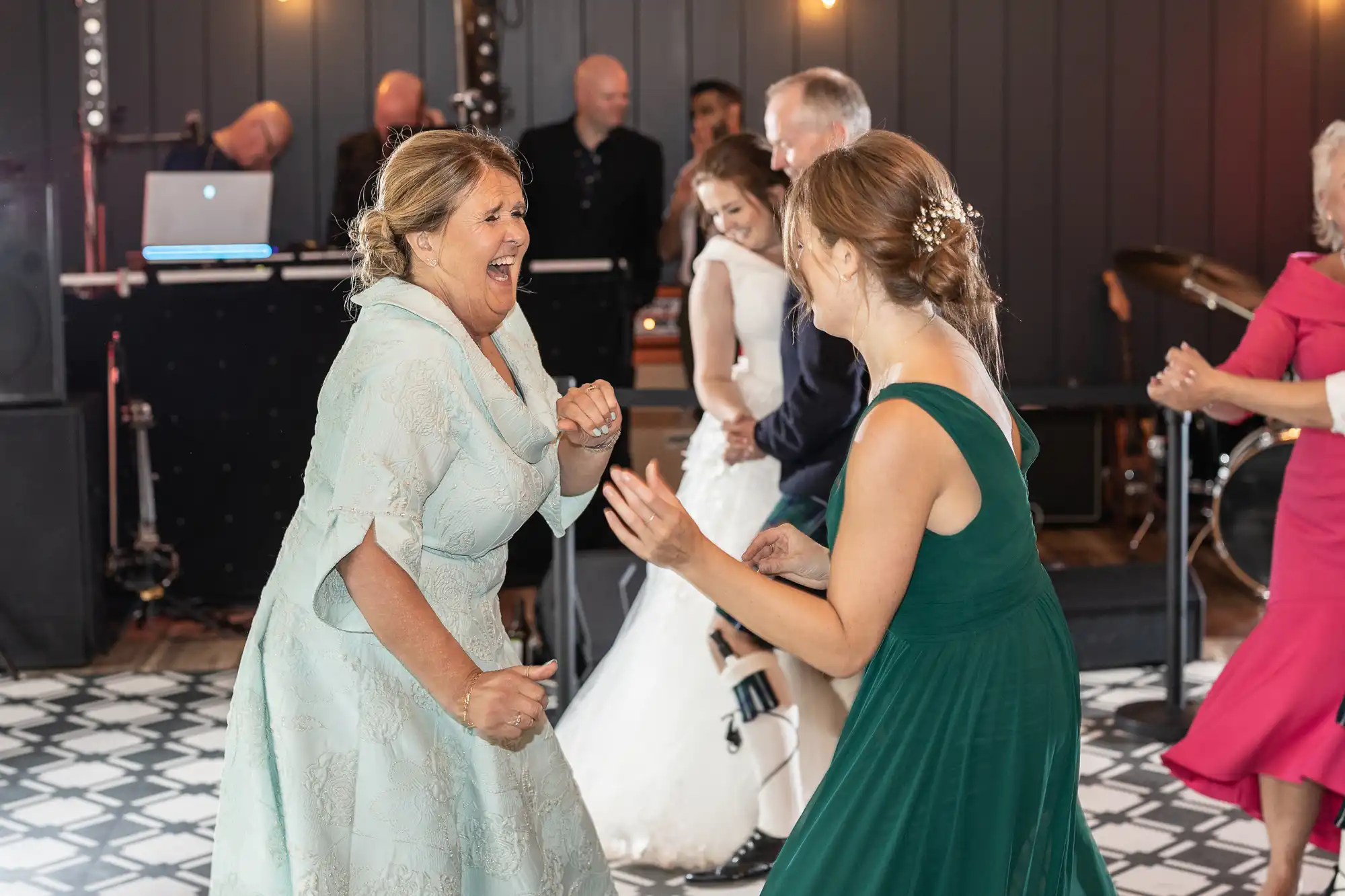 A group of people in formal attire are dancing and smiling in a well-lit room with a band performing in the background. A woman in a mint green dress is laughing energetically.