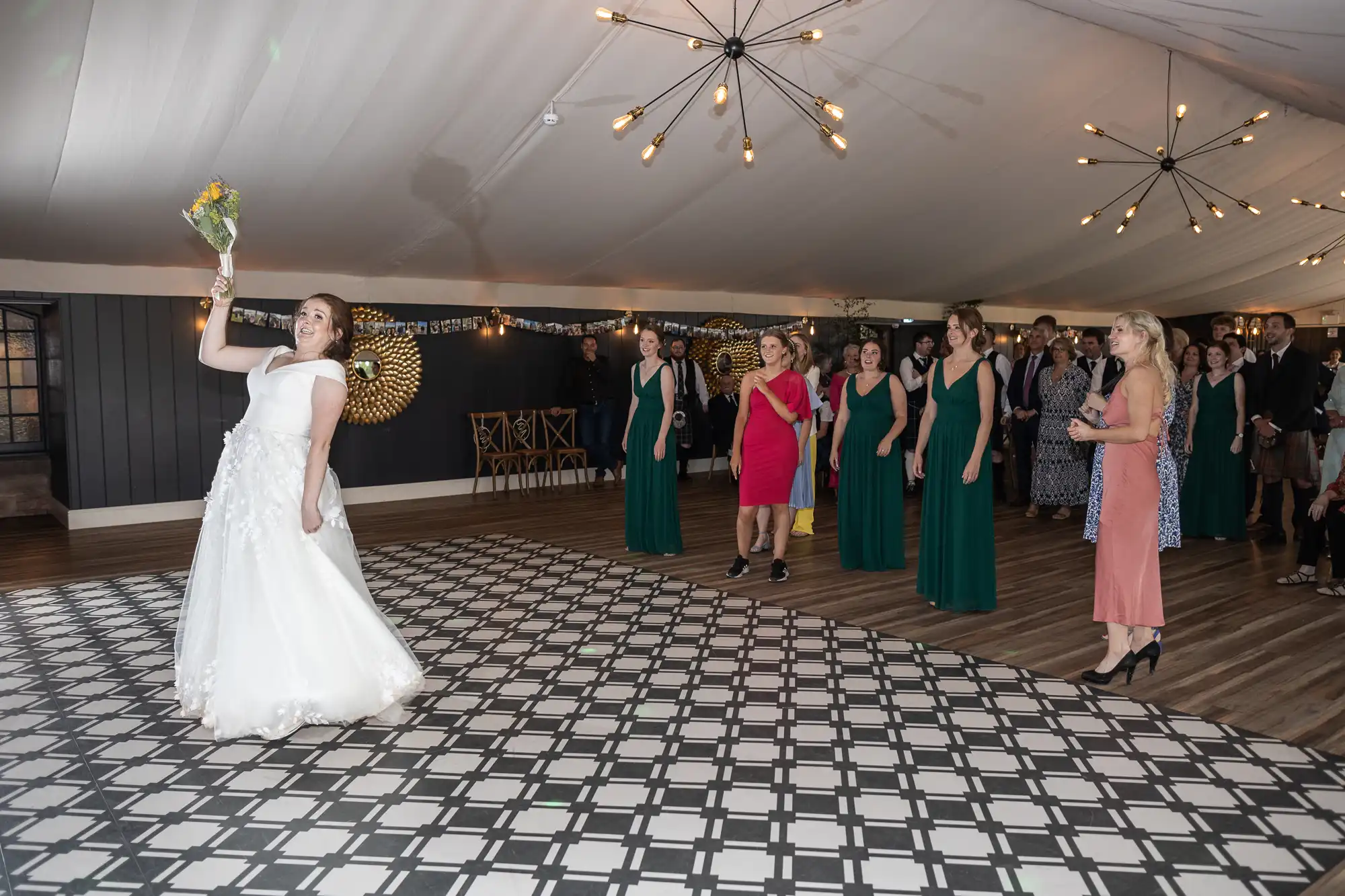 A bride in a white gown prepares to throw a bouquet to a group of women standing behind her on a black-and-white checkered floor.