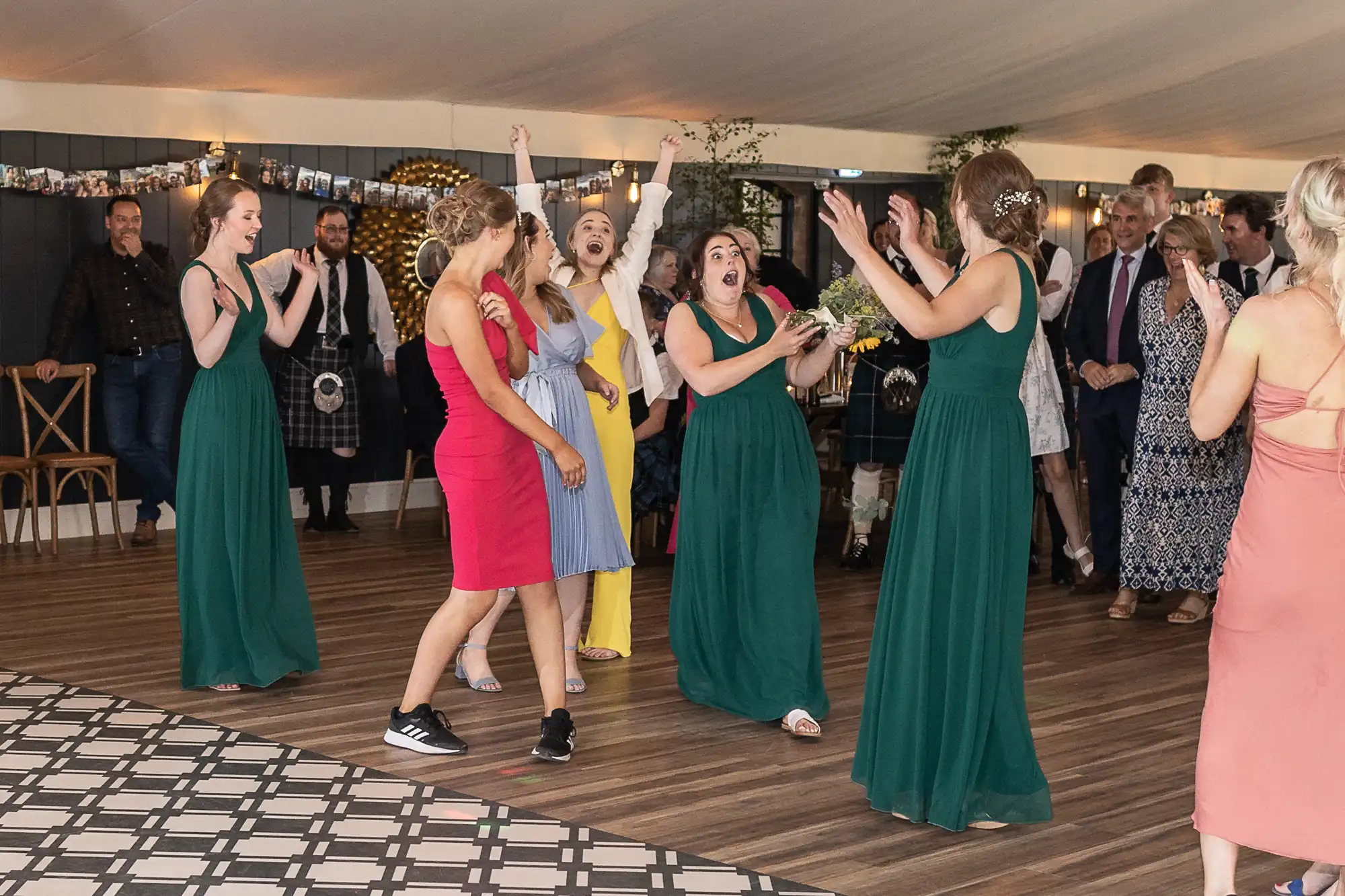 Women are joyfully reacting as one woman in a green dress successfully catches a bouquet at a wedding reception; other guests look on in the background.