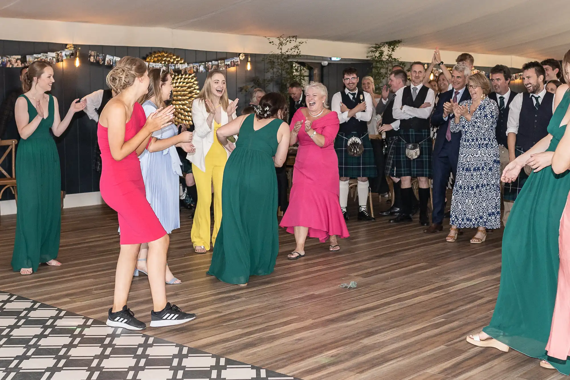 People are joyfully dancing in a room. A group is in the center, some dressed in colorful outfits, while others in kilts and formal attire watch and applaud around them. The atmosphere is lively.