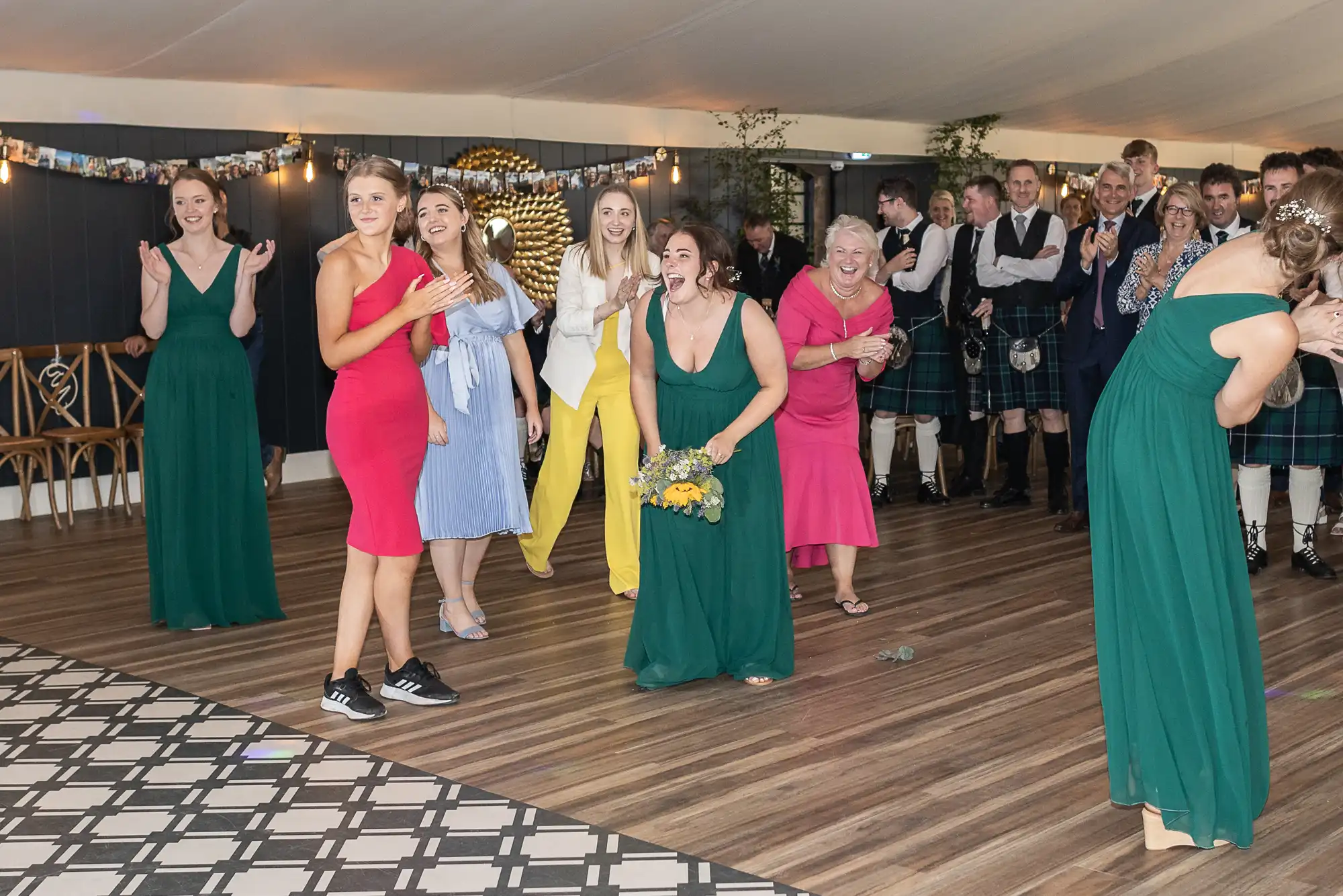 A group of women, in colorful dresses, celebrate as one woman catches a bouquet at a wedding reception. Onlookers, some in formal attire, cheer and smile in the background.