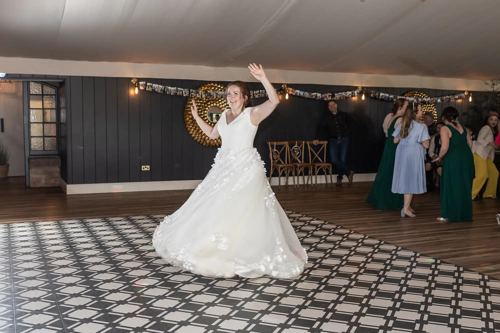 A bride in a white dress dances joyfully on a black-and-white checkered floor while guests watch in the background at a wedding reception.