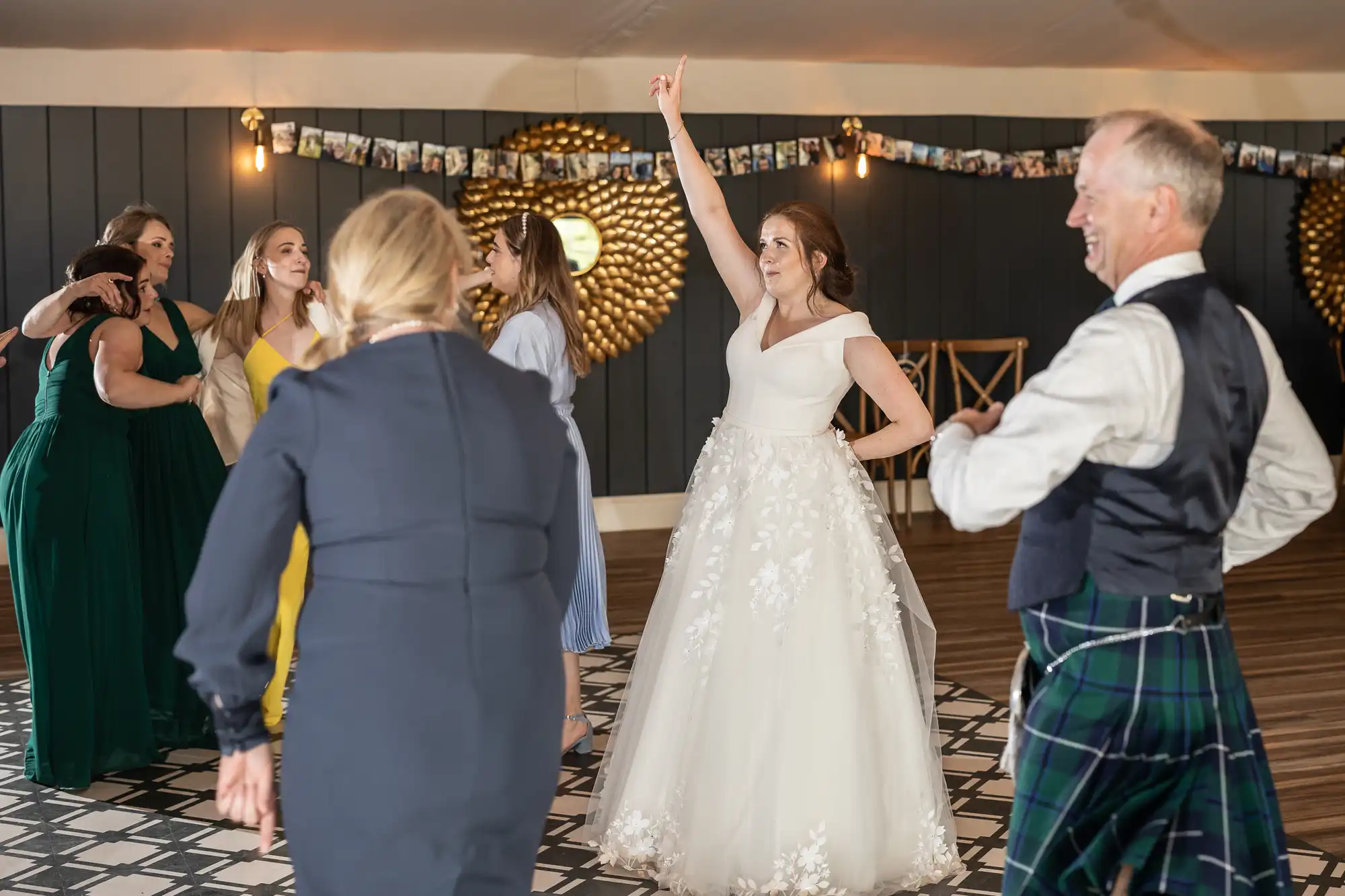 A bride in a white dress and guests, including a man in a kilt, are dancing at a wedding reception with patterned tile floors and dark wall with photo decorations.