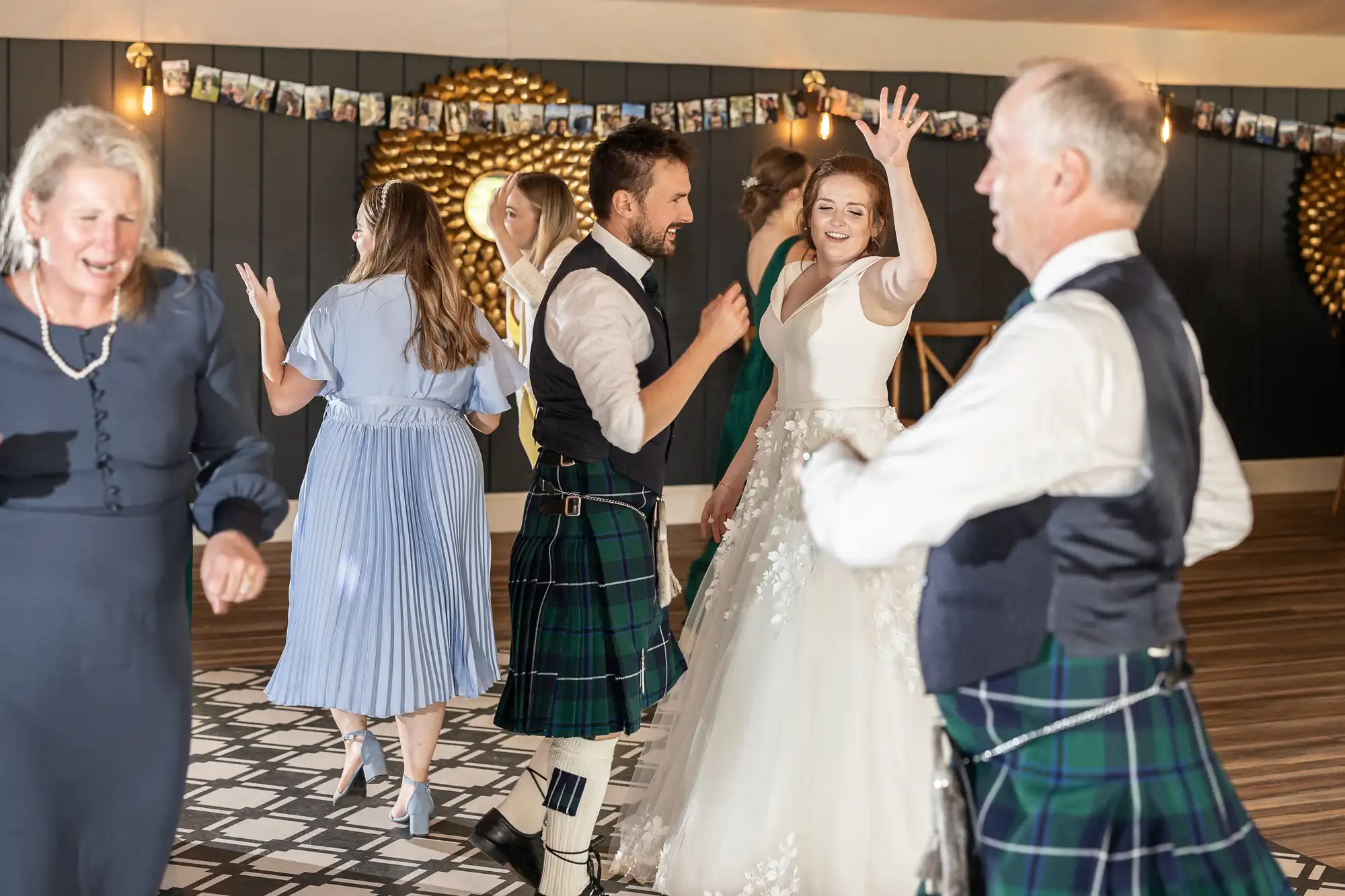 People dressed in formal attire, some in kilts, are dancing and enjoying a celebration in a well-lit room with wooden floor and decor in the background. A bride in a white gown is in the center.