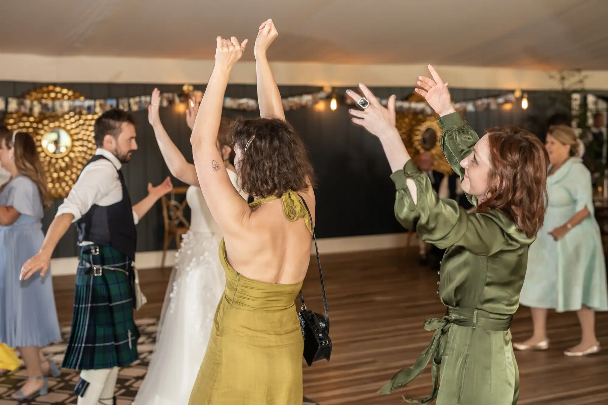 A group of people dancing enthusiastically in a decorated indoor venue, with women in green dresses and a man in a kilt and vest. Some have their arms raised, enjoying the festive atmosphere.