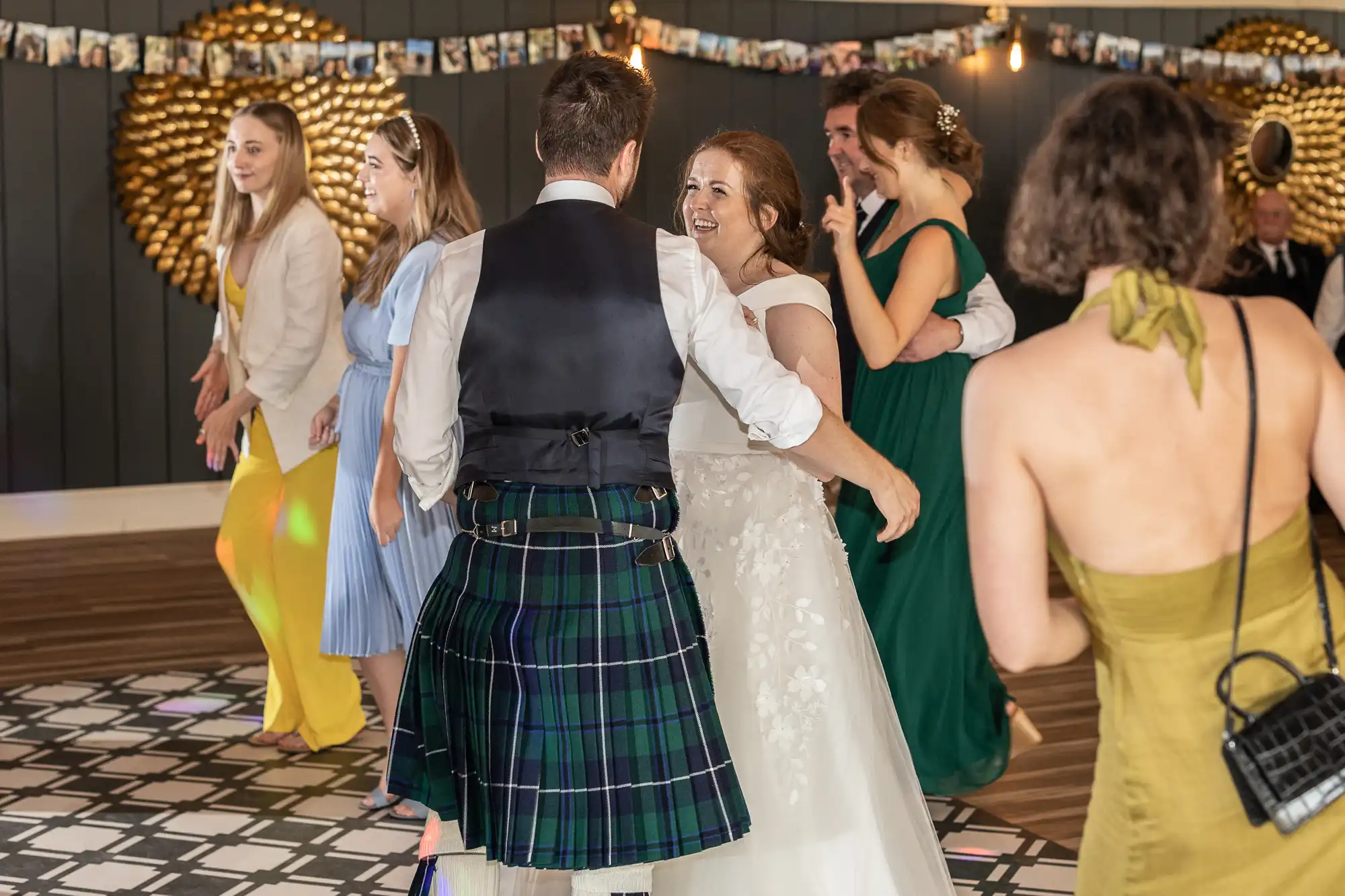 A group of people dancing at a wedding reception. The groom is wearing a kilt and the bride is in a white dress. Other guests in various outfits are visible in the background.