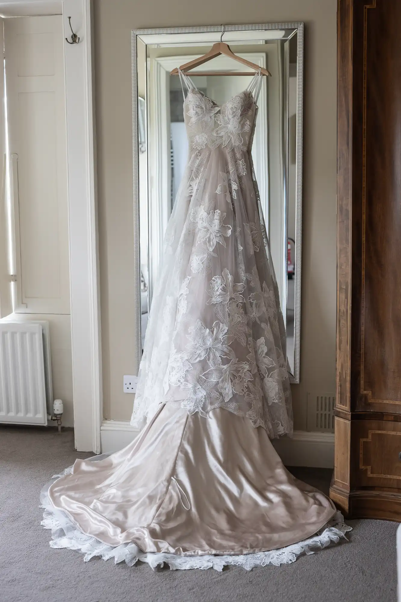 A white floral-patterned wedding dress is hanging on a wooden hanger in front of a tall mirror, with a light-colored satin train spread out on the floor.