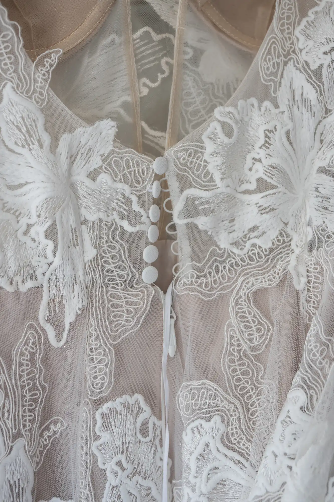 Close-up of the back of a white lace wedding dress, showing intricate floral patterns and a row of small white buttons along the center.