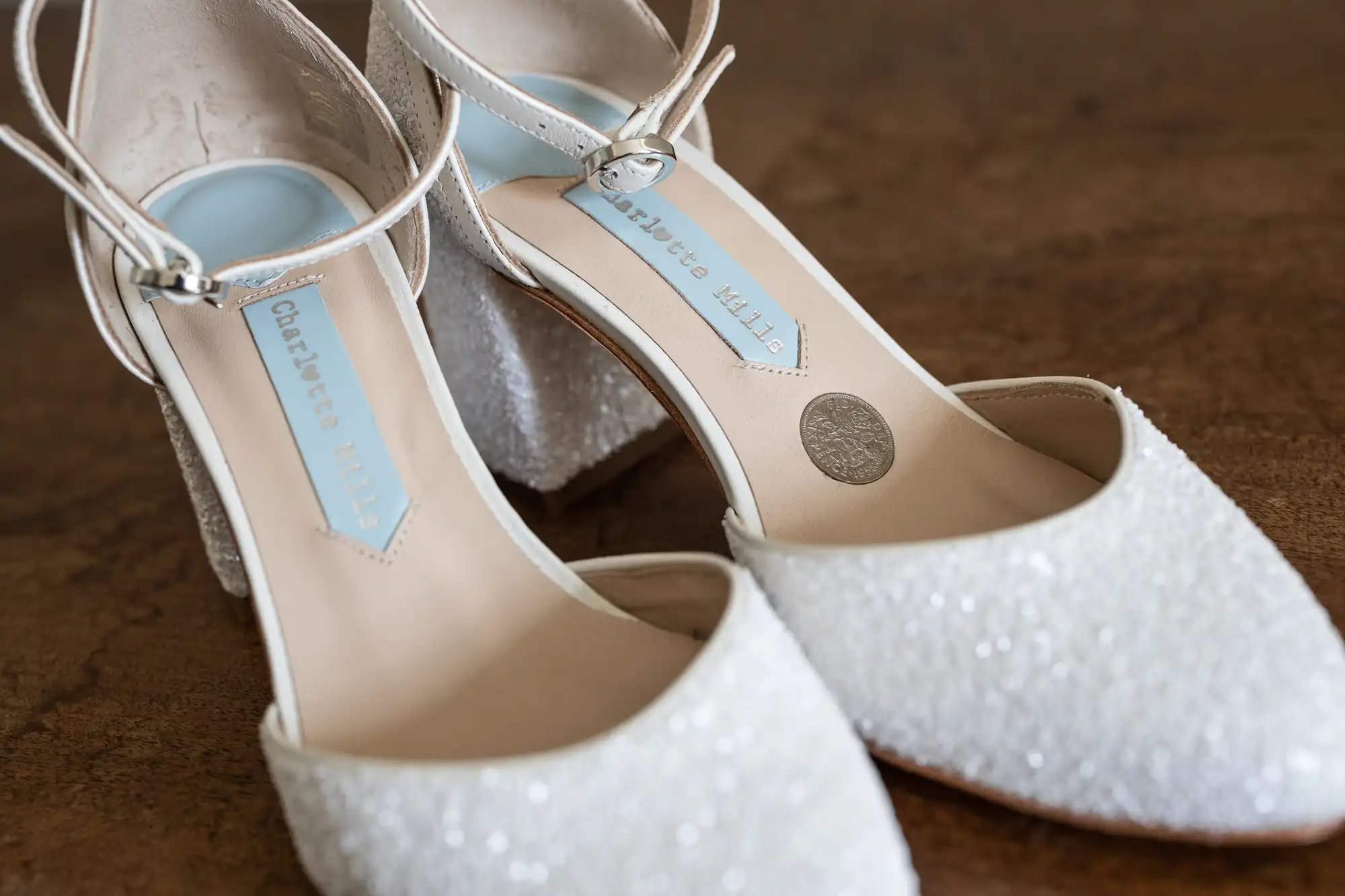 A pair of white sparkly high-heeled shoes with ankle straps, featuring the brand name "Charlotte Mills" on the insole.