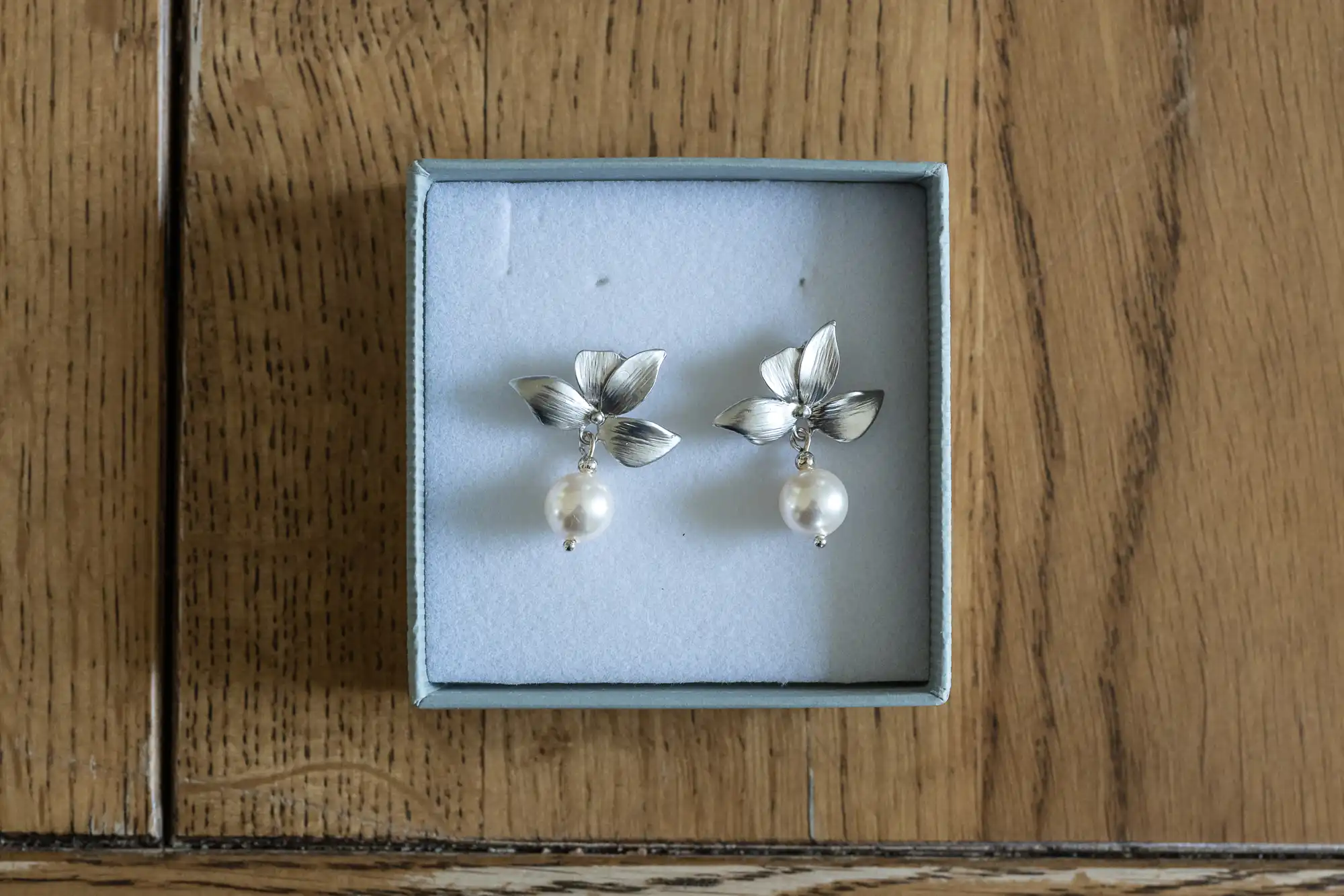 A pair of silver earrings with leaf designs and dangling white pearls placed in an open jewelry box on a wooden surface.