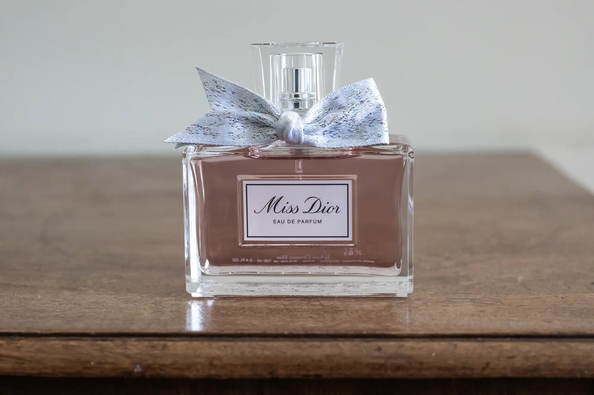 A bottle of Miss Dior Eau de Parfum with a silver-gray bow sits on a wooden surface against a plain background.