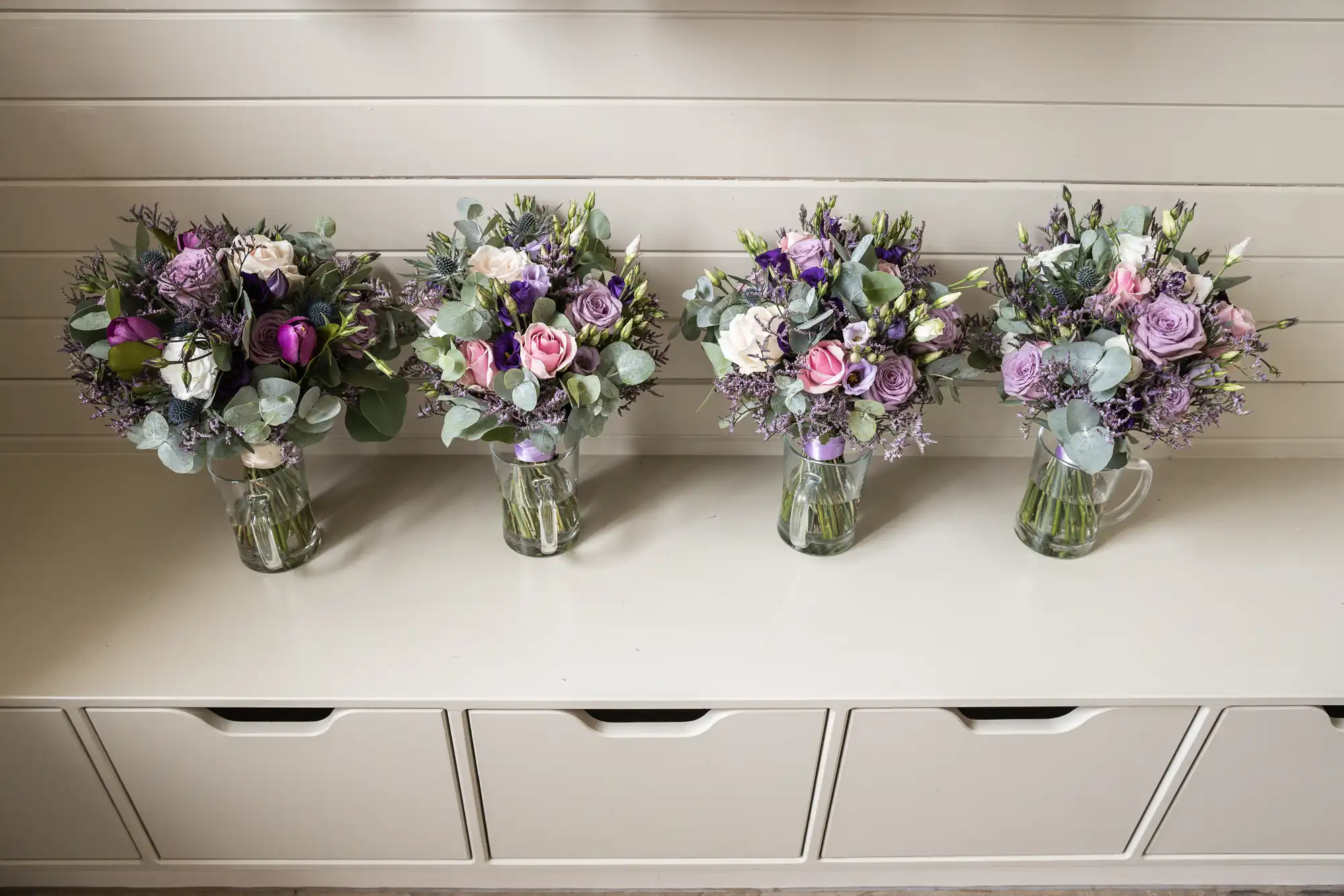 Four floral bouquets in glass pitchers arranged on a white shelf, featuring a mix of purple and white flowers with greenery.