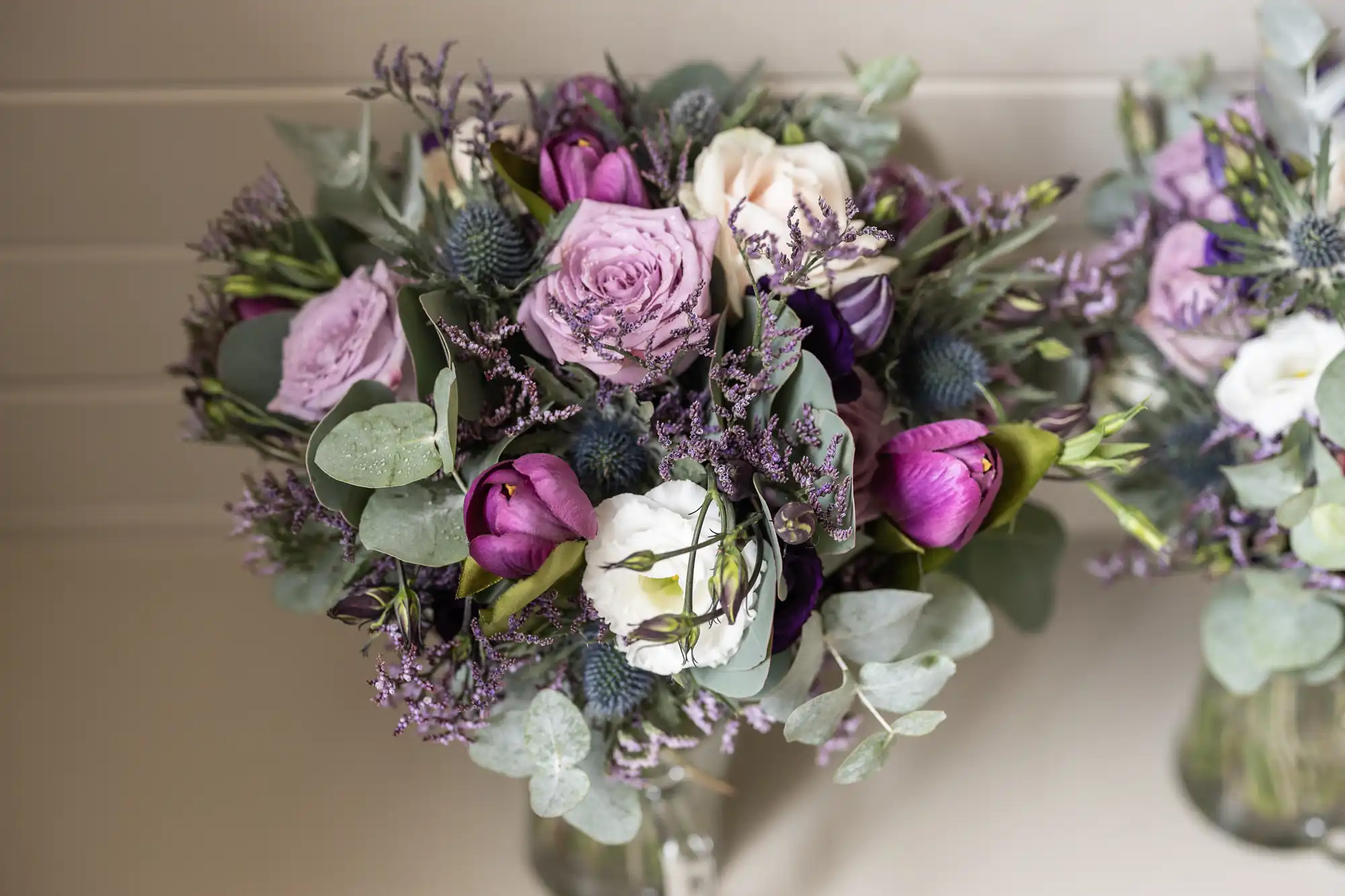 A bouquet featuring purple, white, and pink flowers mixed with greenery and purple filler flowers in a vase, placed against a light-colored background.