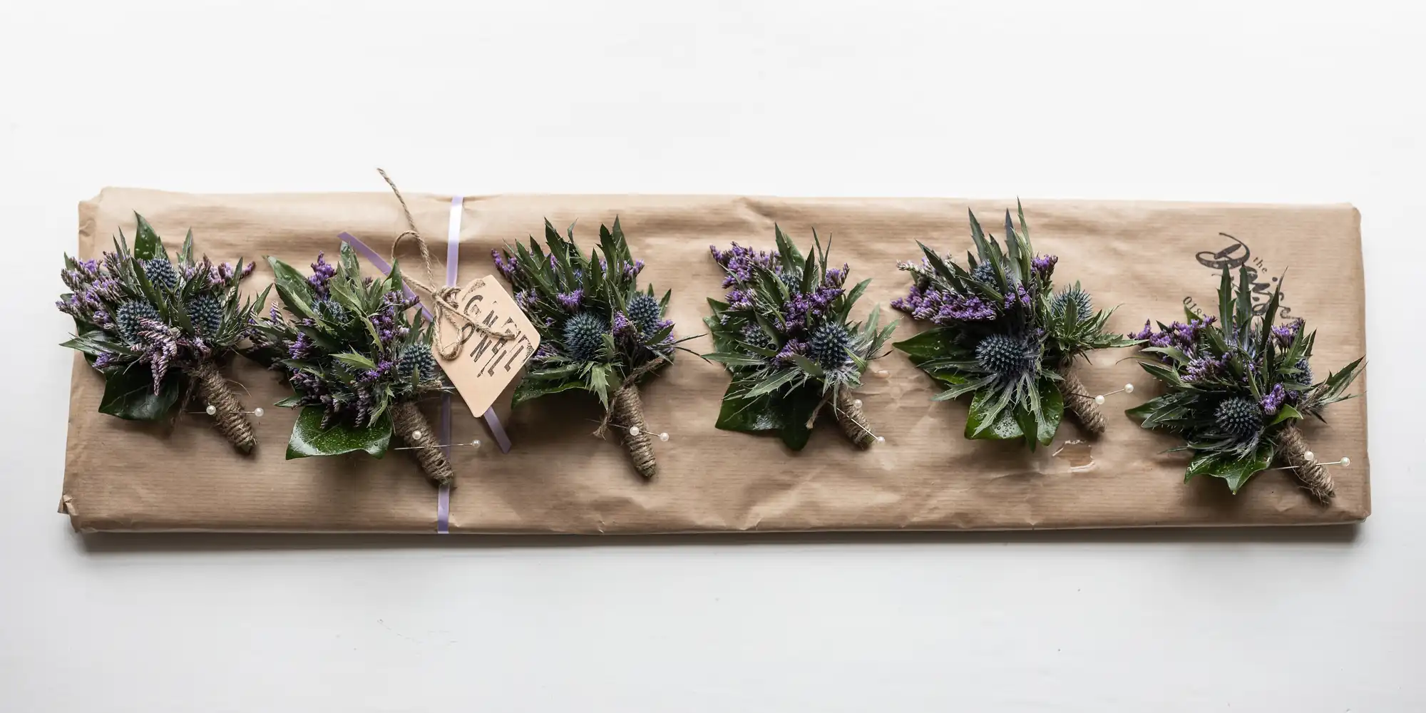 Six boutonnières made of thistle and purple flowers wrapped in brown paper, arranged in a line.