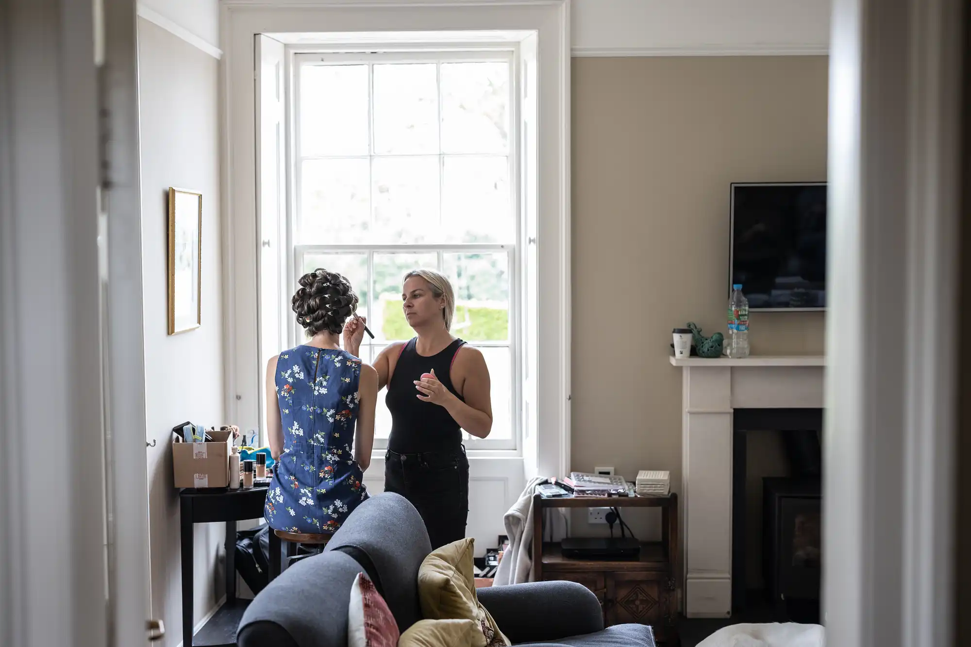 A person is getting their makeup done by a makeup artist in a brightly lit living room with a large window. The room has a couch, fireplace, and TV.