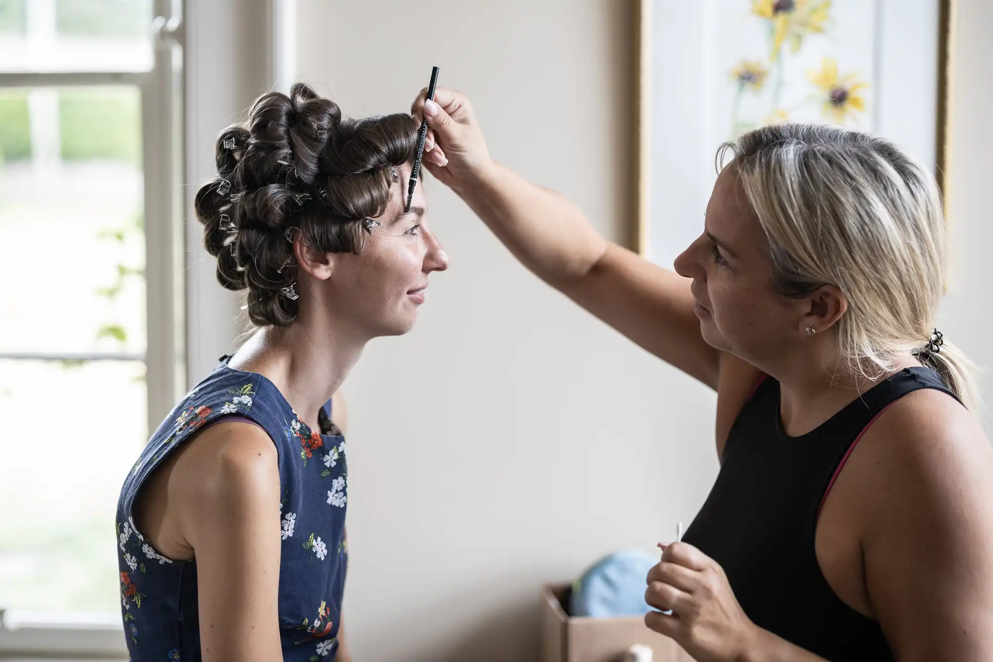 A woman with curled hair sits as another woman applies makeup with a brush in a well-lit room.