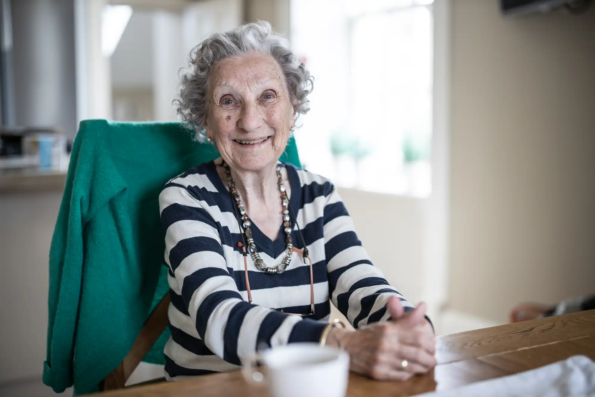 An elderly woman with curly gray hair and wearing a black and white striped shirt sits smiling at a wooden table. A green blanket is draped over her chair. A cup and saucer are on the table.