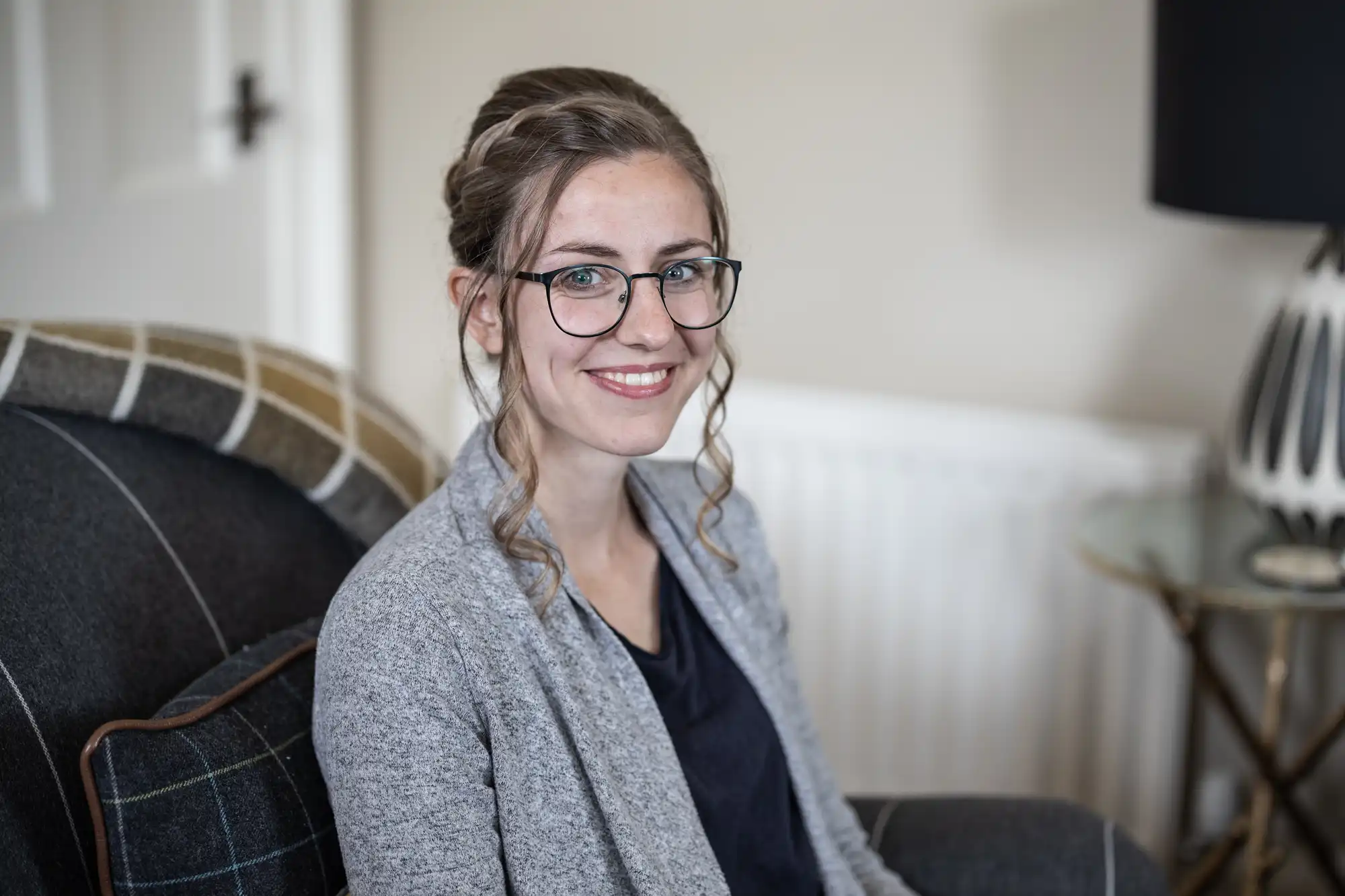 A person with glasses and a gray cardigan sits in a living room, smiling at the camera.