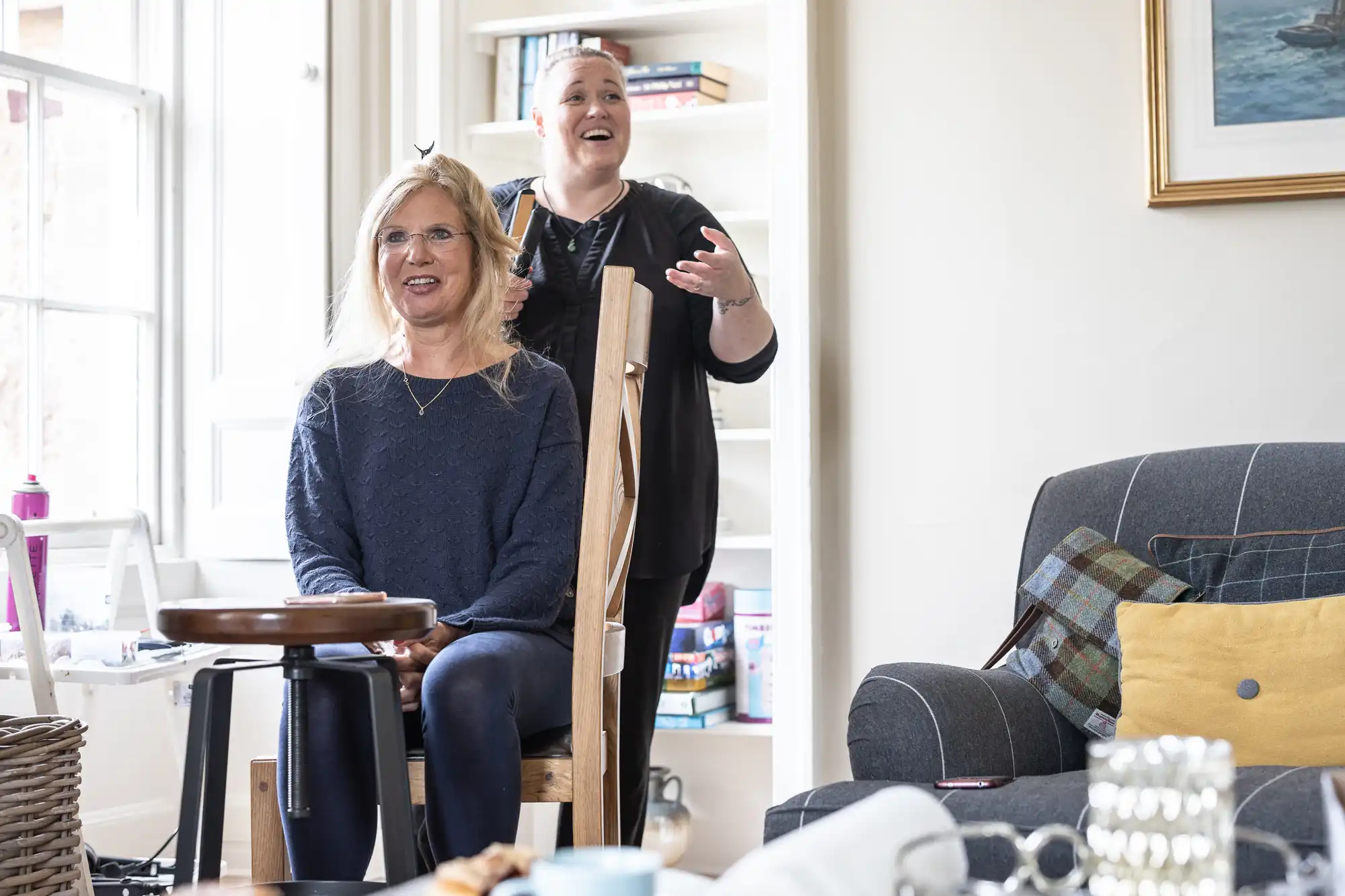 A smiling woman sits on a stool, having her hair styled by another woman in a cozy living room with bookshelves and a window in the background.