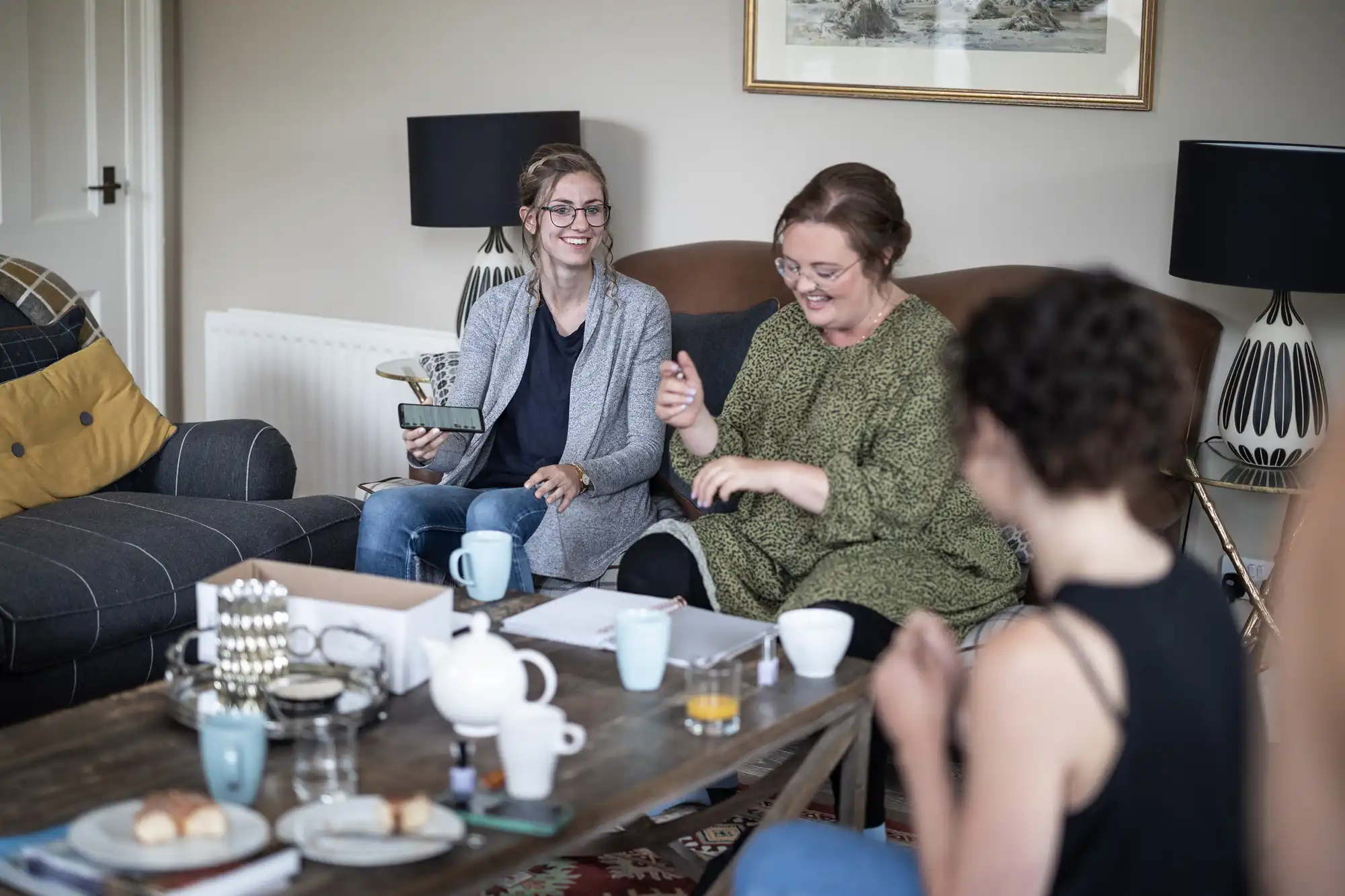 Three people are sitting around a coffee table with cups and snacks, chatting and smiling in a living room. One person holds a smartphone while another gesticulates animatedly.