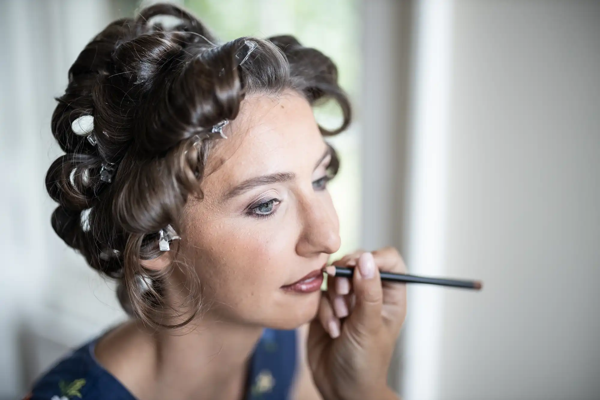 A person with hair in rollers is having makeup applied to their lips with a pencil by another person.