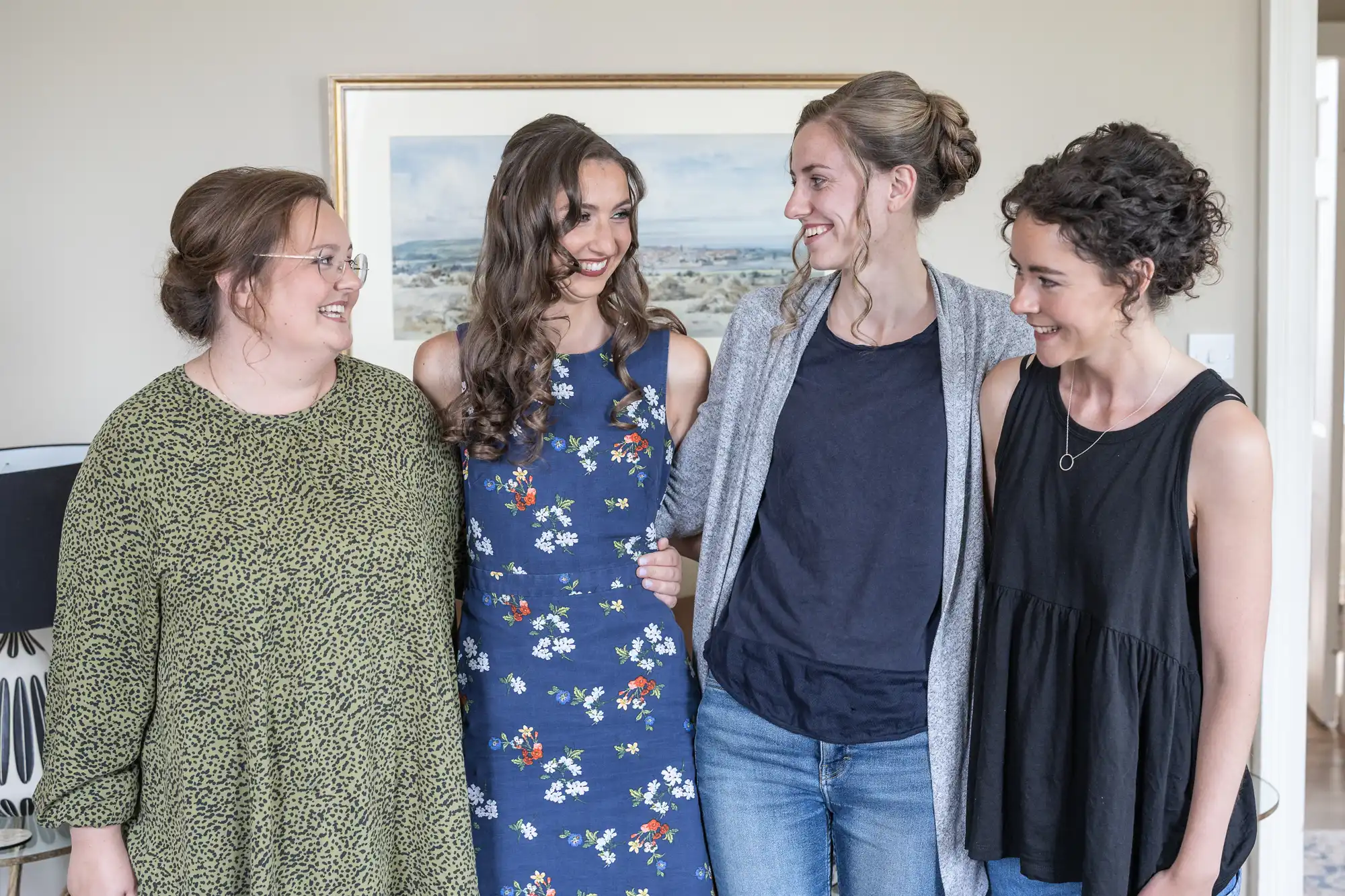 Four women smiling and standing together indoors, each dressed in casual attire. A framed picture hangs on the wall behind them.