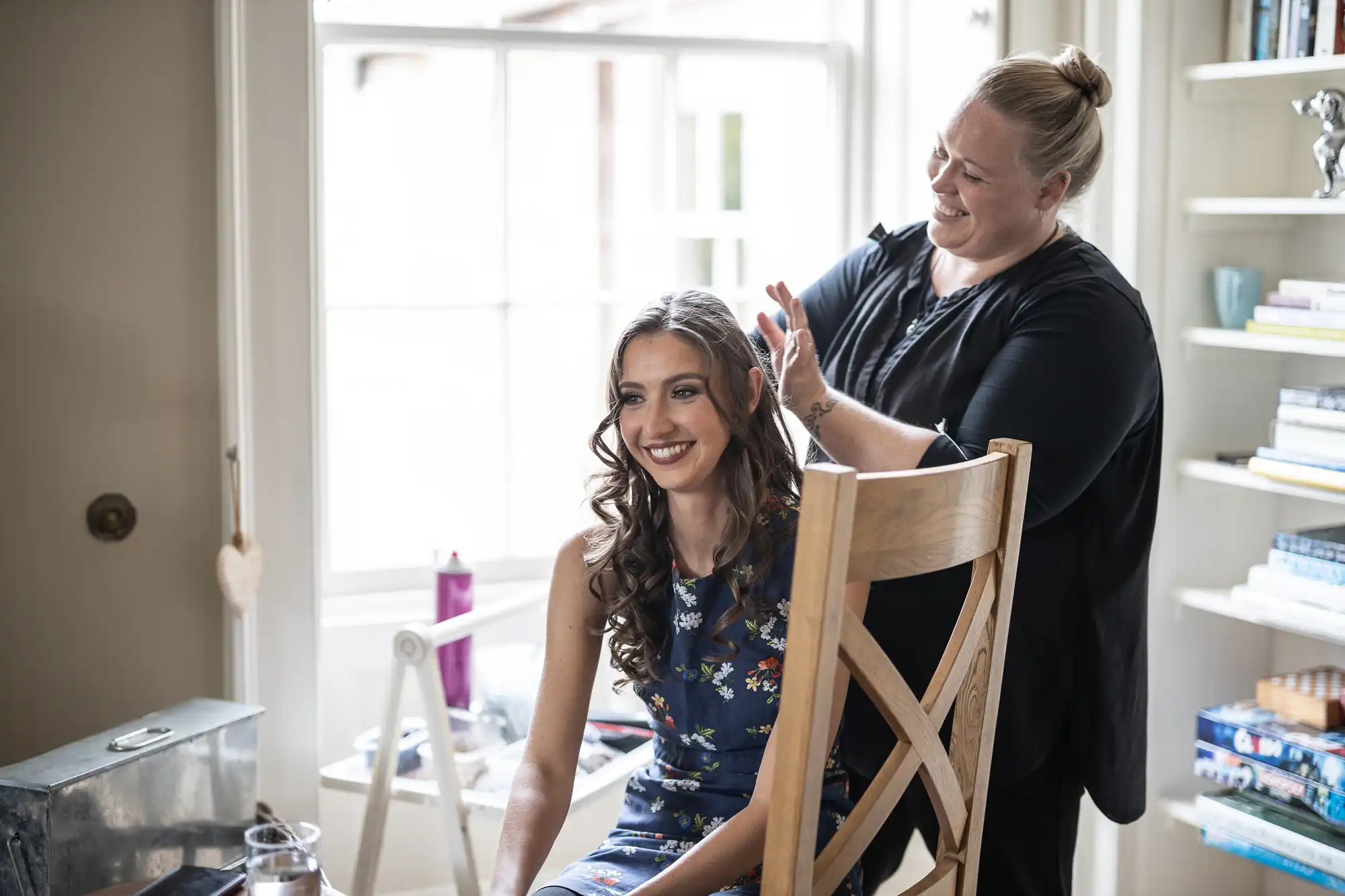 A woman with long hair sits smiling in a chair while another woman stands behind her, styling her hair. They are in a well-lit room with a window and bookshelves.