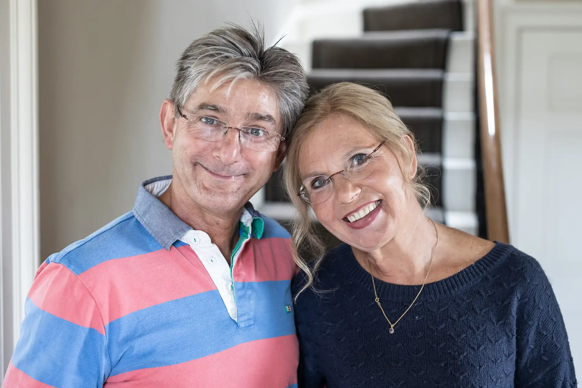 A man and woman, both smiling, stand close together indoors. The woman rests her head on the man's shoulder. A staircase is visible in the background.