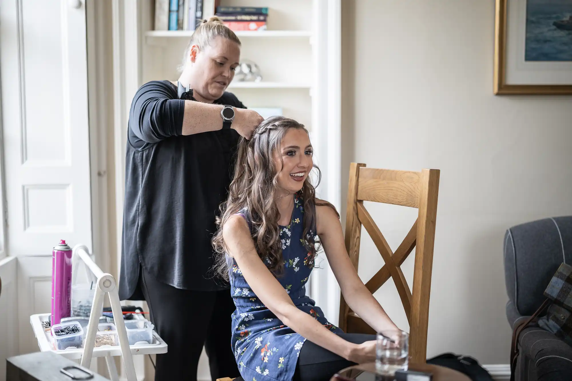 A woman styles another woman's hair in a bright room. The seated woman is smiling and wearing a floral dress. A glass of water is on the table beside her.