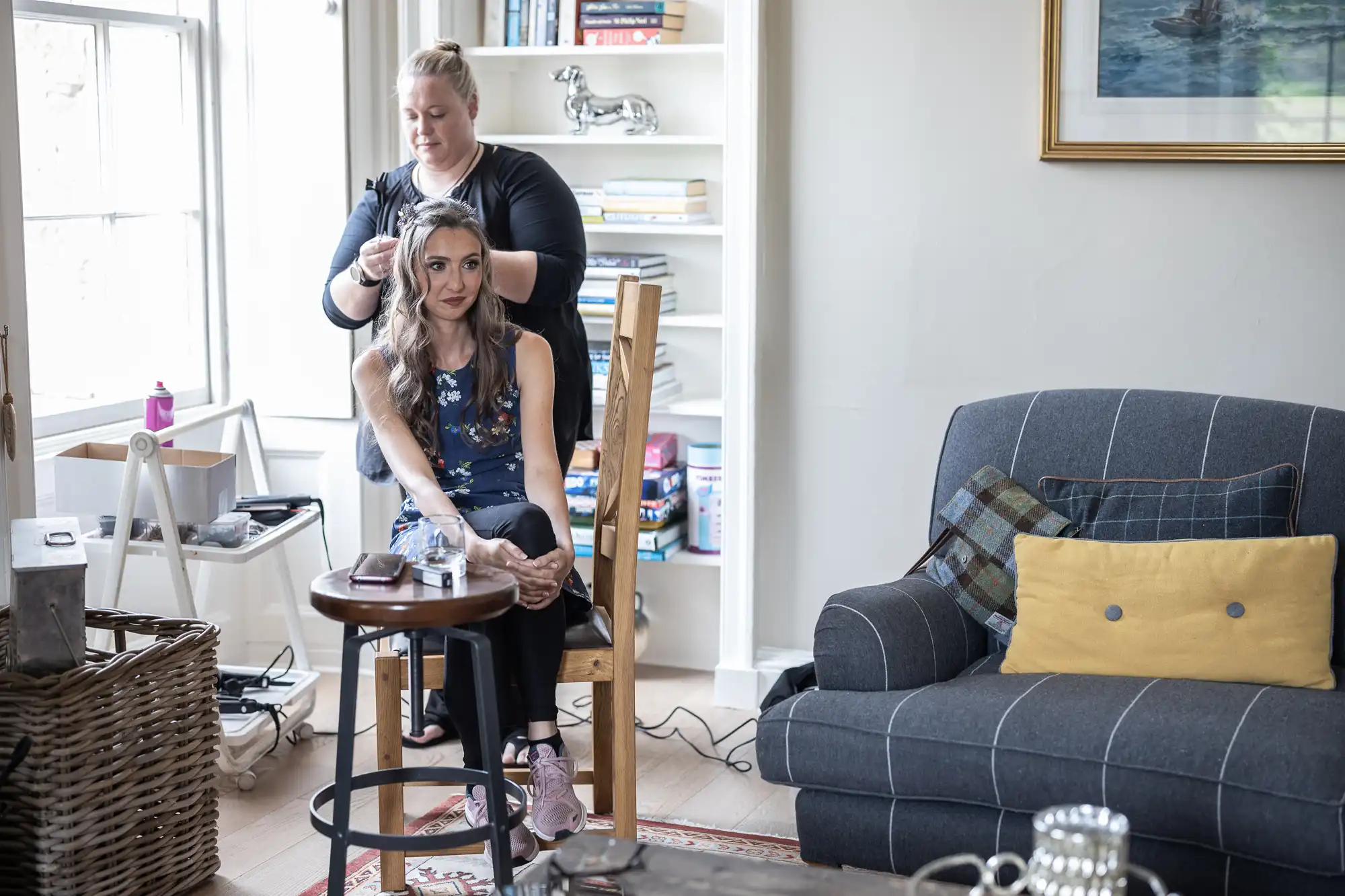 A woman is seated on a wooden chair in a living room while another woman styles her hair. A sofa, shelves with books, and a framed picture are visible in the background.