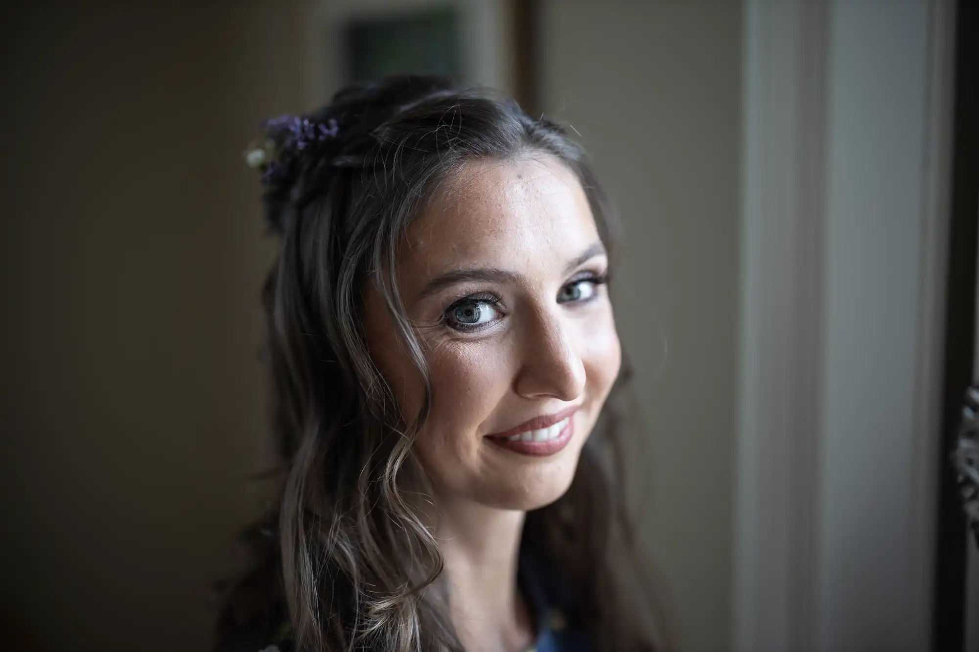 A woman with brown wavy hair and blue eyes is smiling while looking at the camera in an indoor setting. She has flowers in her hair.