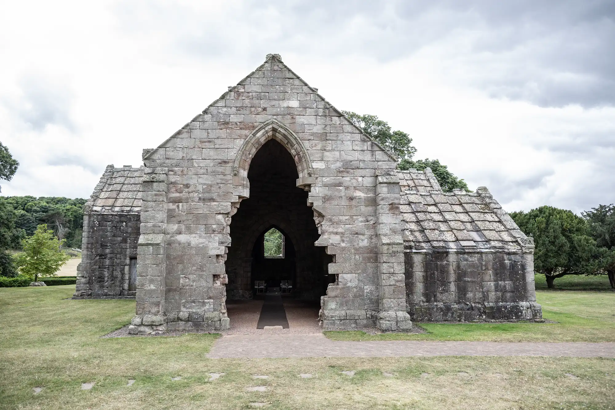 A stone church ruin with an arched entrance, partially collapsed walls, and a grassy foreground under a cloudy sky.