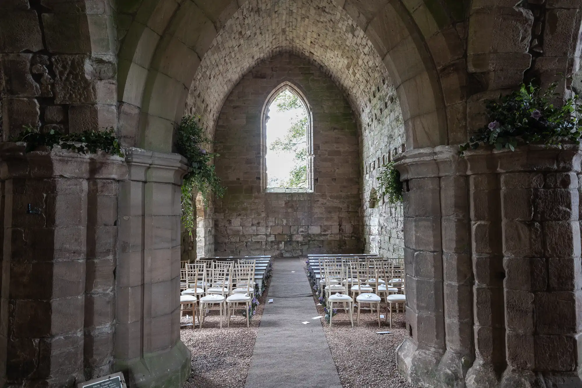 Interior of an old stone chapel with rows of white chairs arranged for a ceremony, facing toward a tall, narrow arched window at the far end.