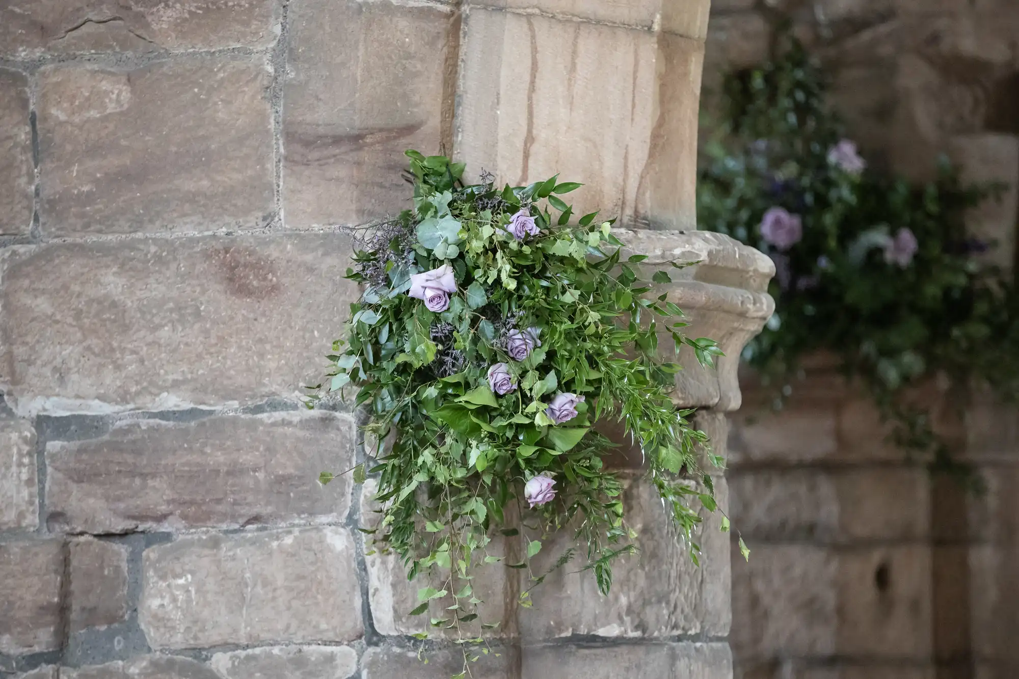 A stone pillar adorned with a bouquet of purple and green foliage. Another similar bouquet is in the background, attached to a stone wall.