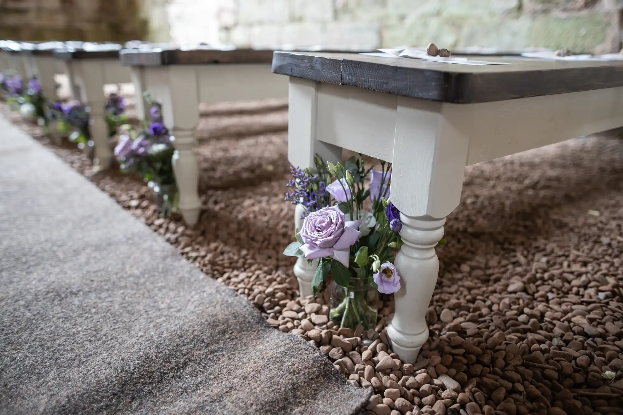 Close-up of benches lined up on a gravel pathway, each adorned with small arrangements of purple and white flowers.