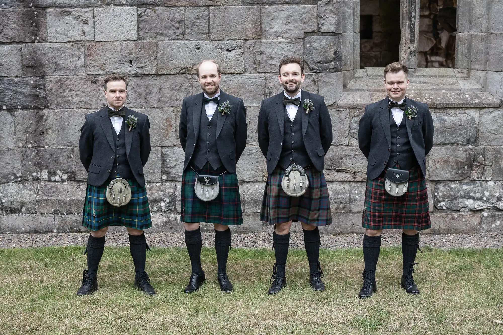 Four men wearing traditional Scottish kilts and jackets stand in a row in front of a stone wall.