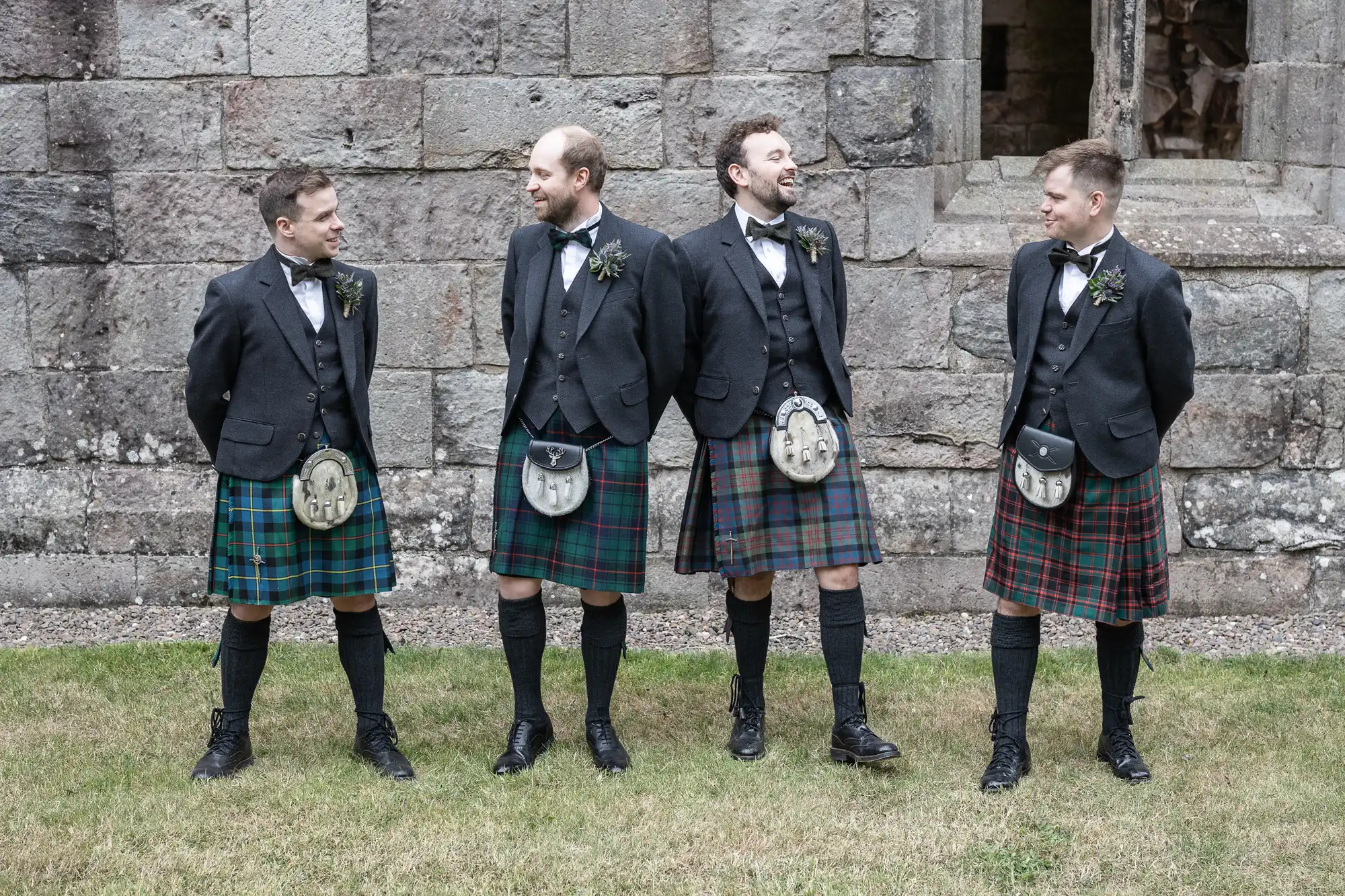 Four men wearing kilts, jackets, and sporrans stand in a row in front of a stone building, smiling and looking at each other.