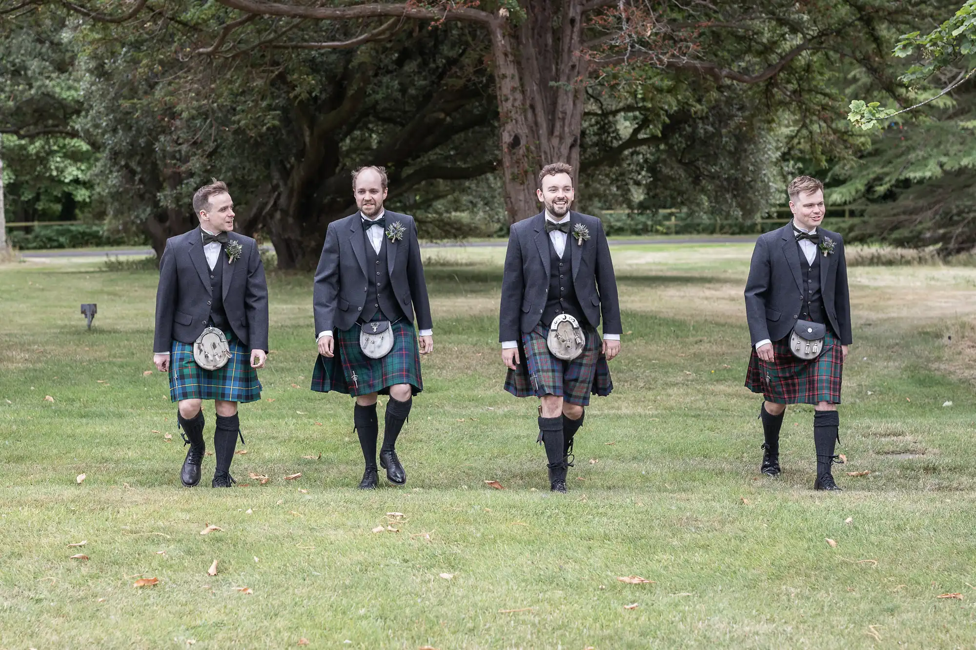 Four men, dressed in traditional Scottish kilts and formal jackets, walk together on a grassy area with trees in the background.