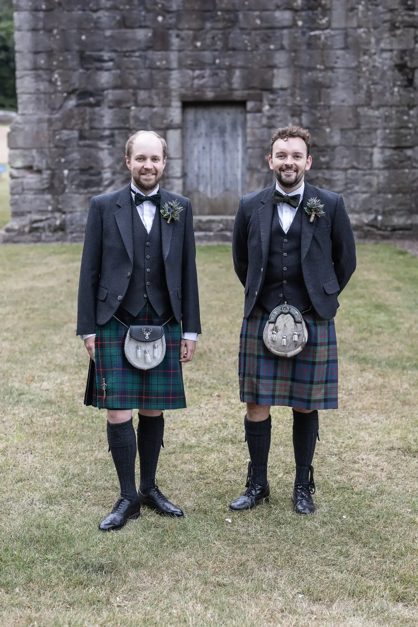 Two men in formal attire, wearing traditional kilts and sporrans, stand side by side on a grassy area with a stone building in the background.