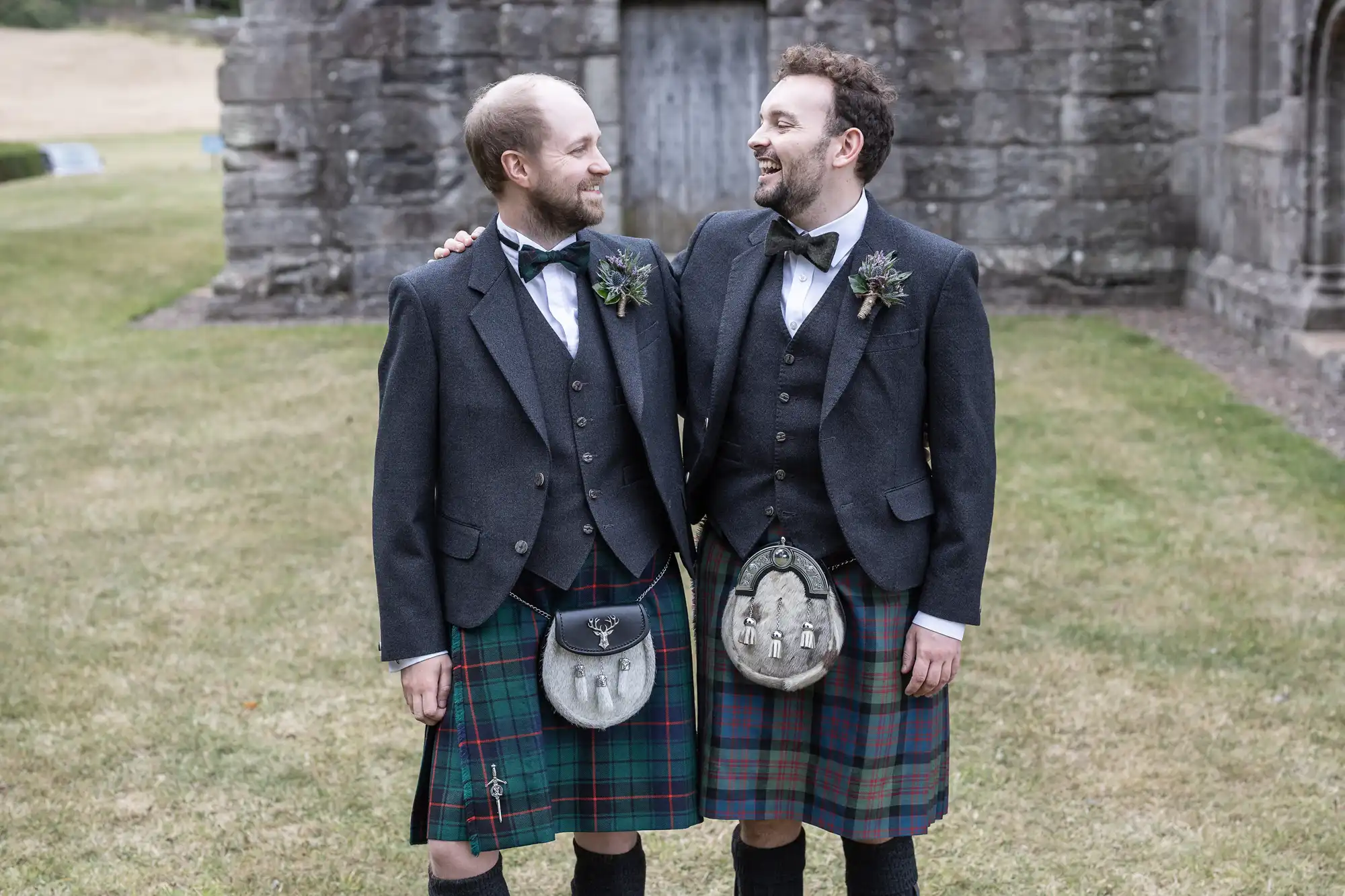 Two men in traditional Scottish attire, including kilts and sporrans, stand close together outside a stone building, smiling at each other.