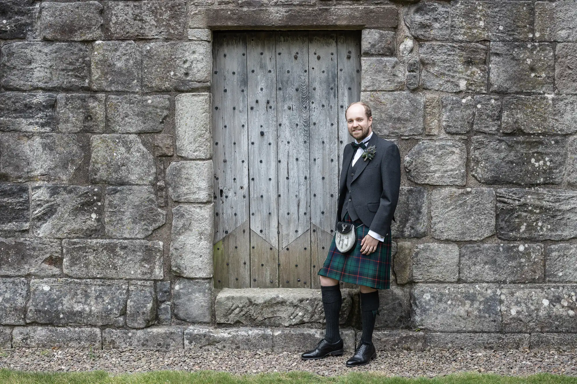 A man in formal attire, including a kilt, stands against a stone wall beside a wooden door, smiling at the camera.