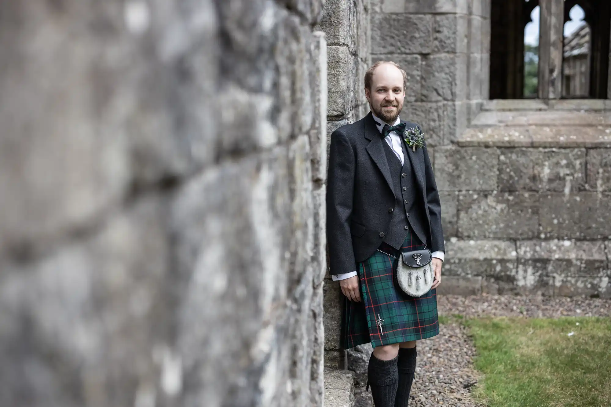 A bald man with a beard stands outside by a stone wall, wearing traditional Scottish attire including a black jacket, a green and red kilt, a sporran, and knee-high socks. There is grass and a stone building nearby.