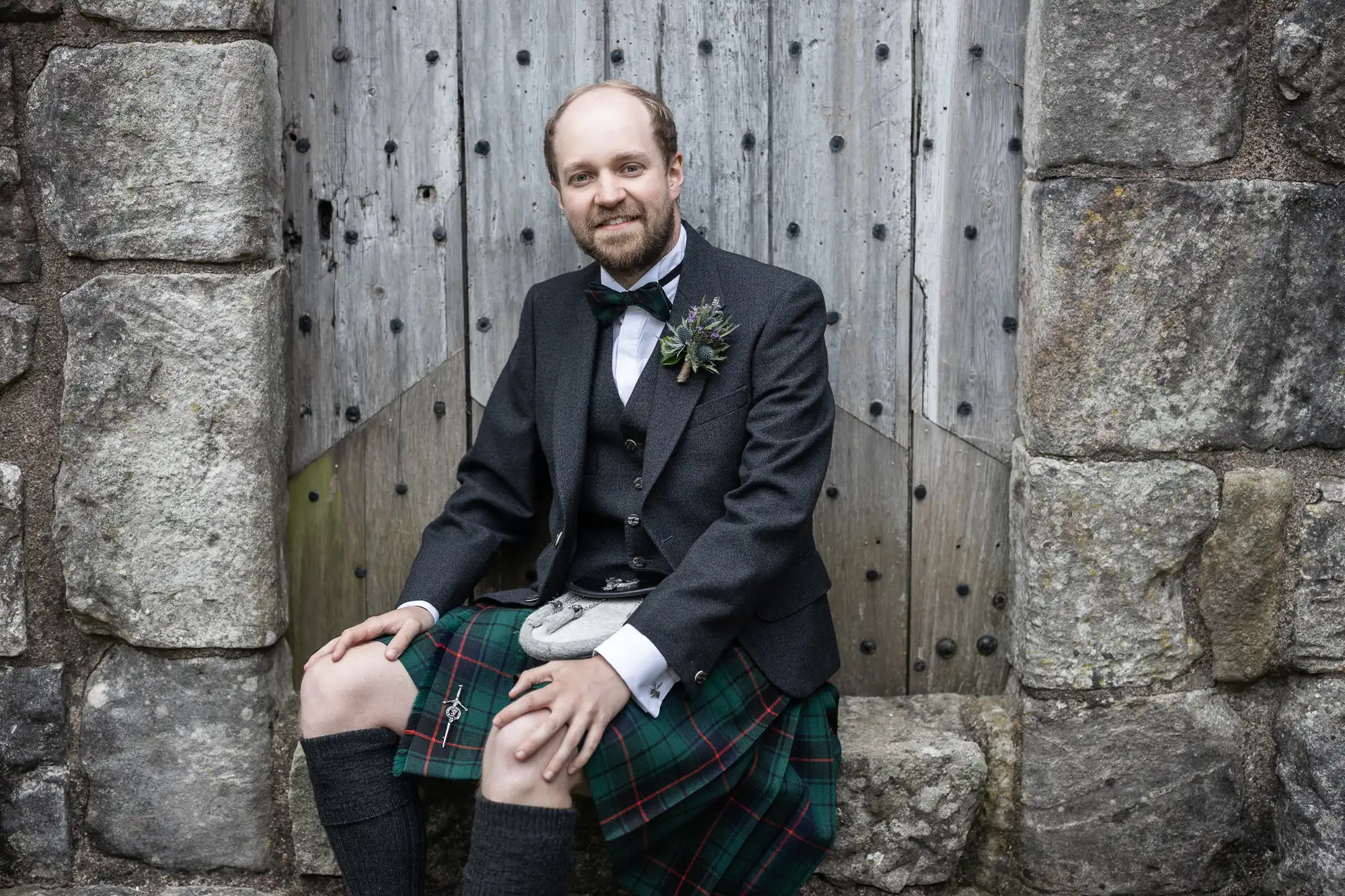 A man in formal Scottish attire, including a kilt and jacket, sits on a stone step against a weathered wooden door with metal studs.