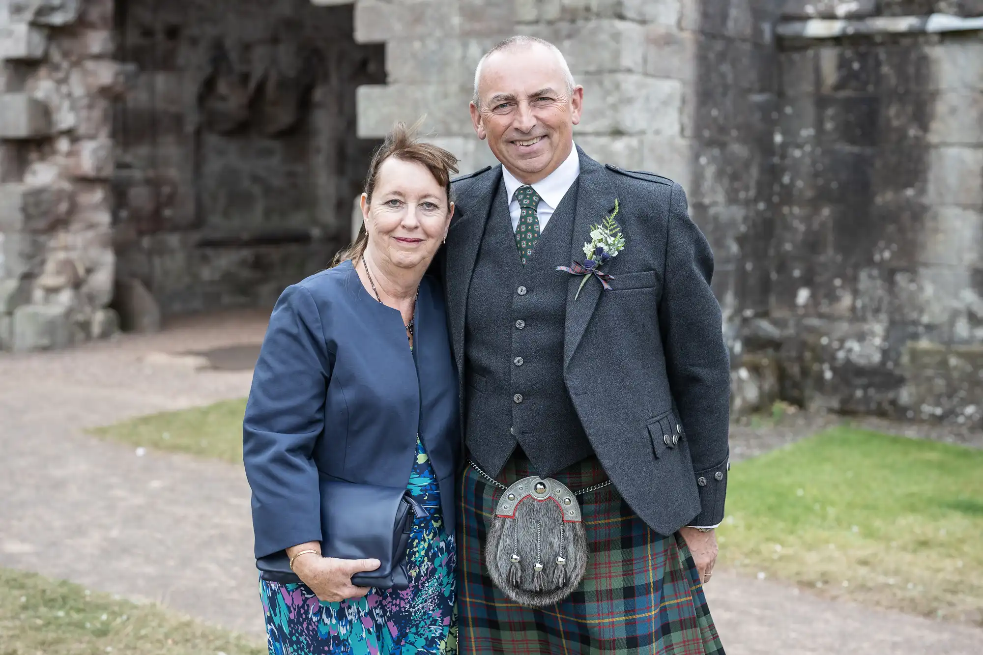 A man and a woman standing outside beside a stone building. The man is wearing traditional Scottish attire, and the woman is dressed in a blue outfit. They are both smiling at the camera.