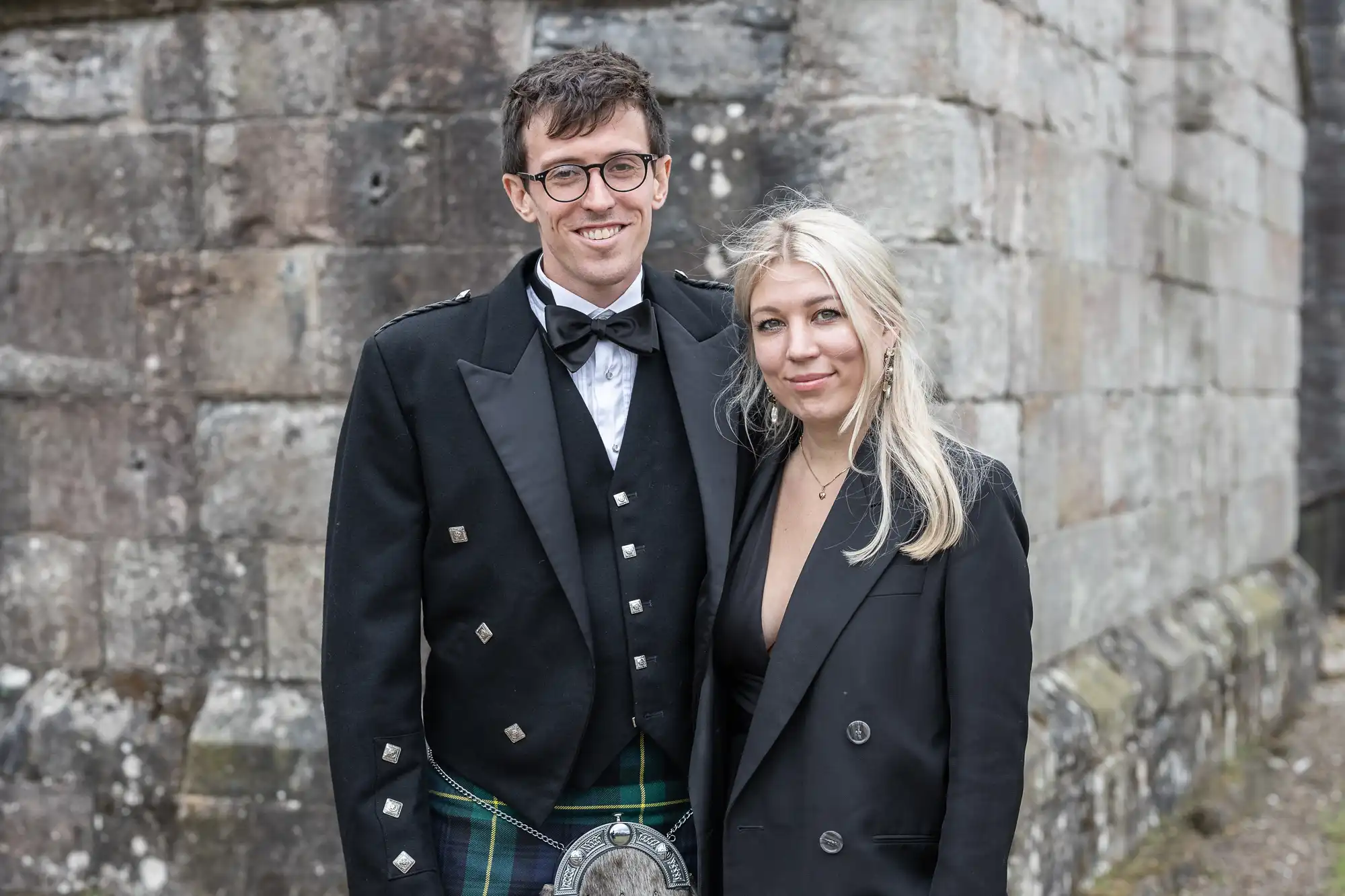 A man and a woman posing for a photo against a stone wall. The man is wearing glasses and traditional Scottish attire, while the woman is wearing a black outfit.